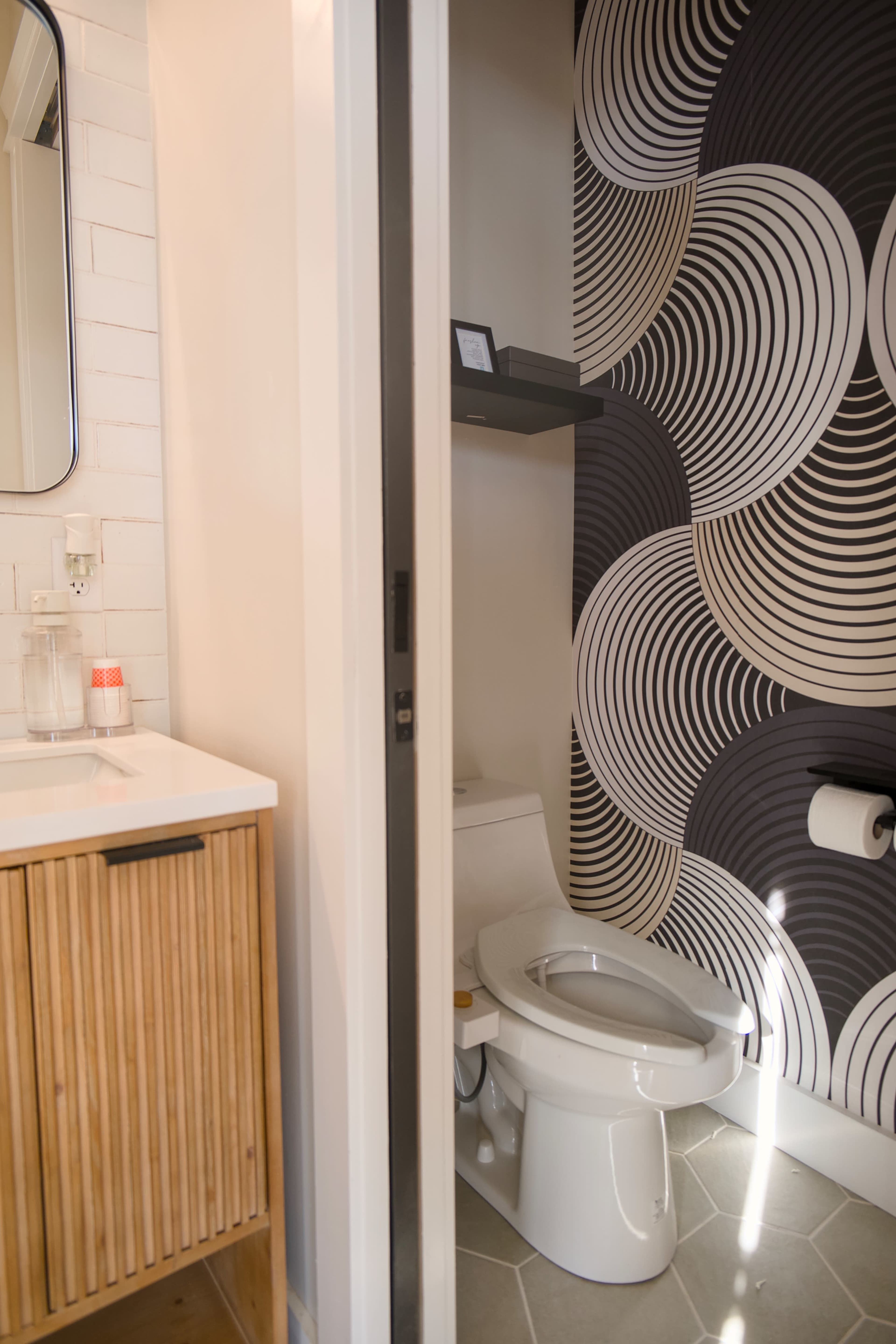The image shows a modern bathroom with a separate toilet area featuring a patterned accent wall and a wooden vanity with a sink.
