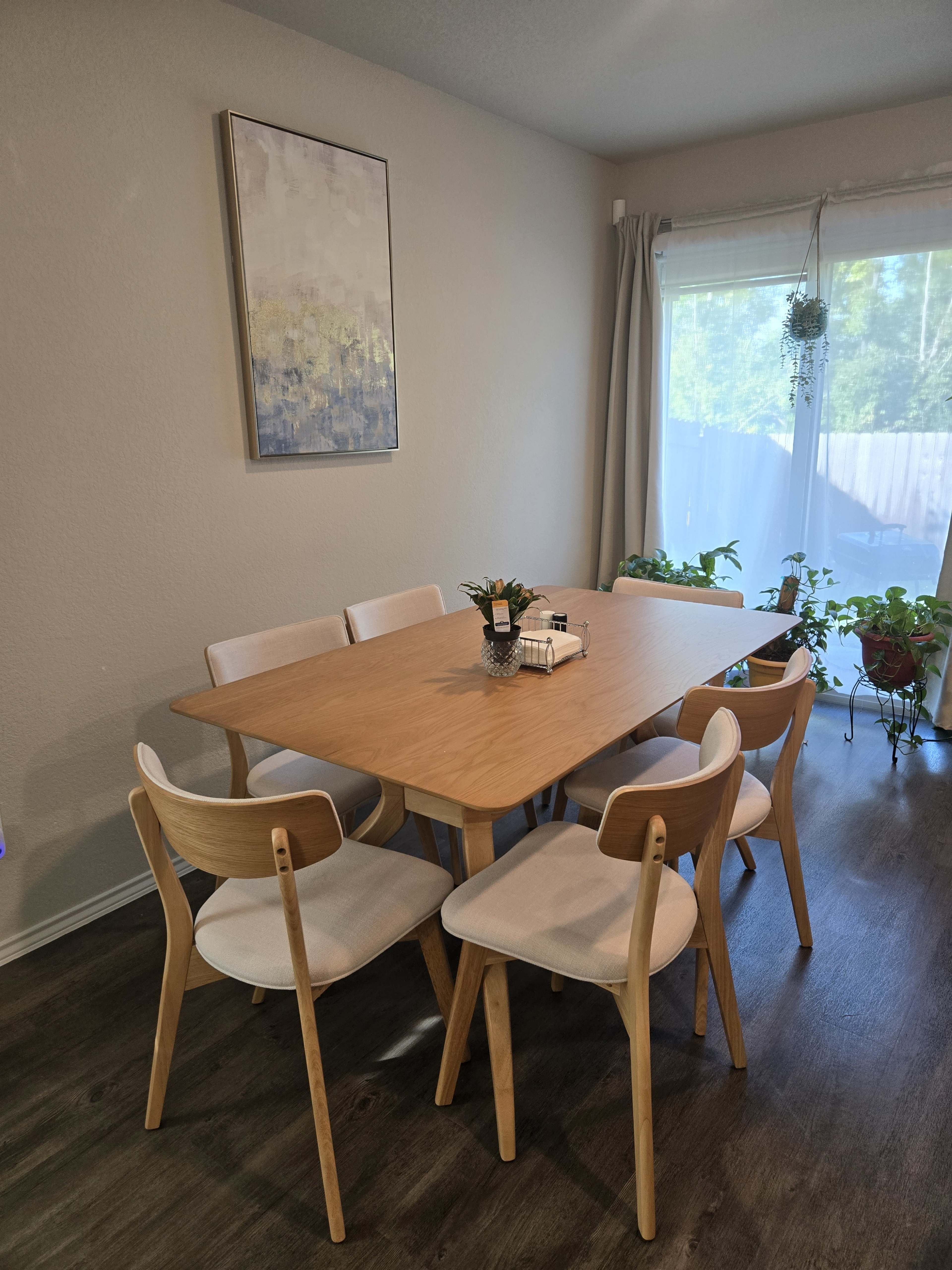 A wooden dining table surrounded by six chairs is positioned in a well-lit room with a window featuring plants and decor.
