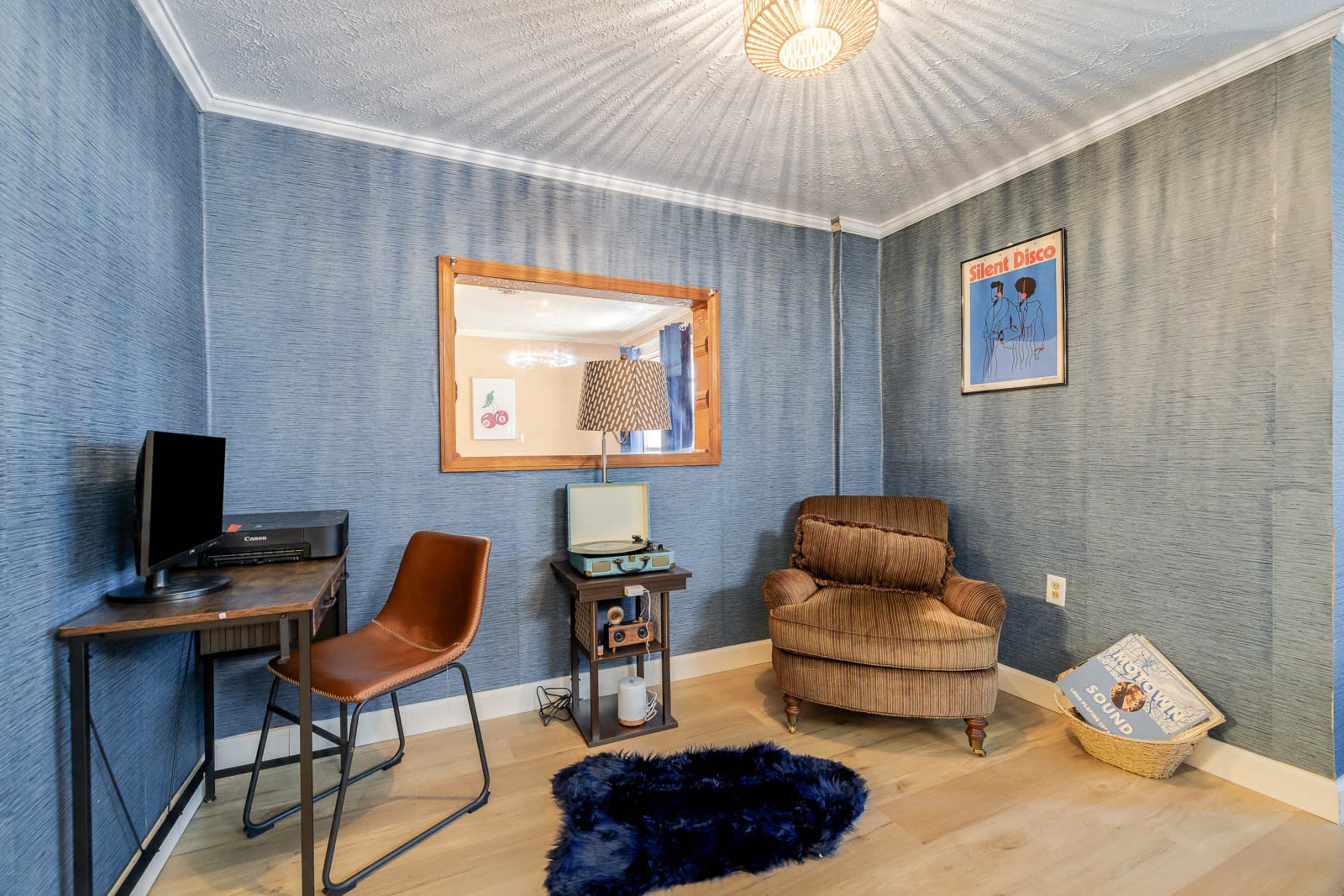 The image shows a small room with blue textured walls, featuring a desk with a computer, a vintage television on a stand, a comfortable brown armchair, and a blue rug on the floor.