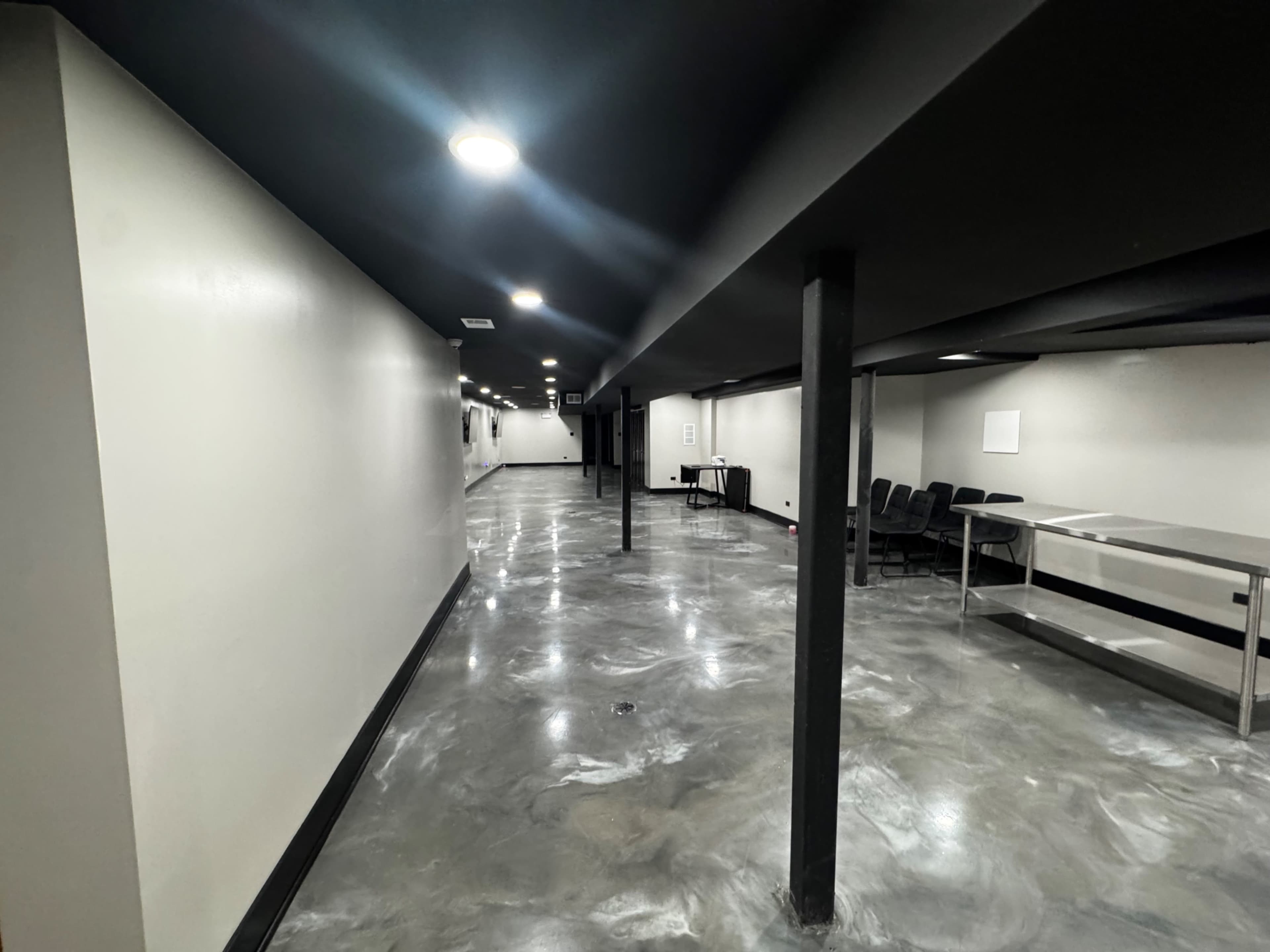 The image shows a long, narrow corridor with polished concrete flooring, overhead lighting, and black pillars, leading to an area with chairs and a table on the right side.