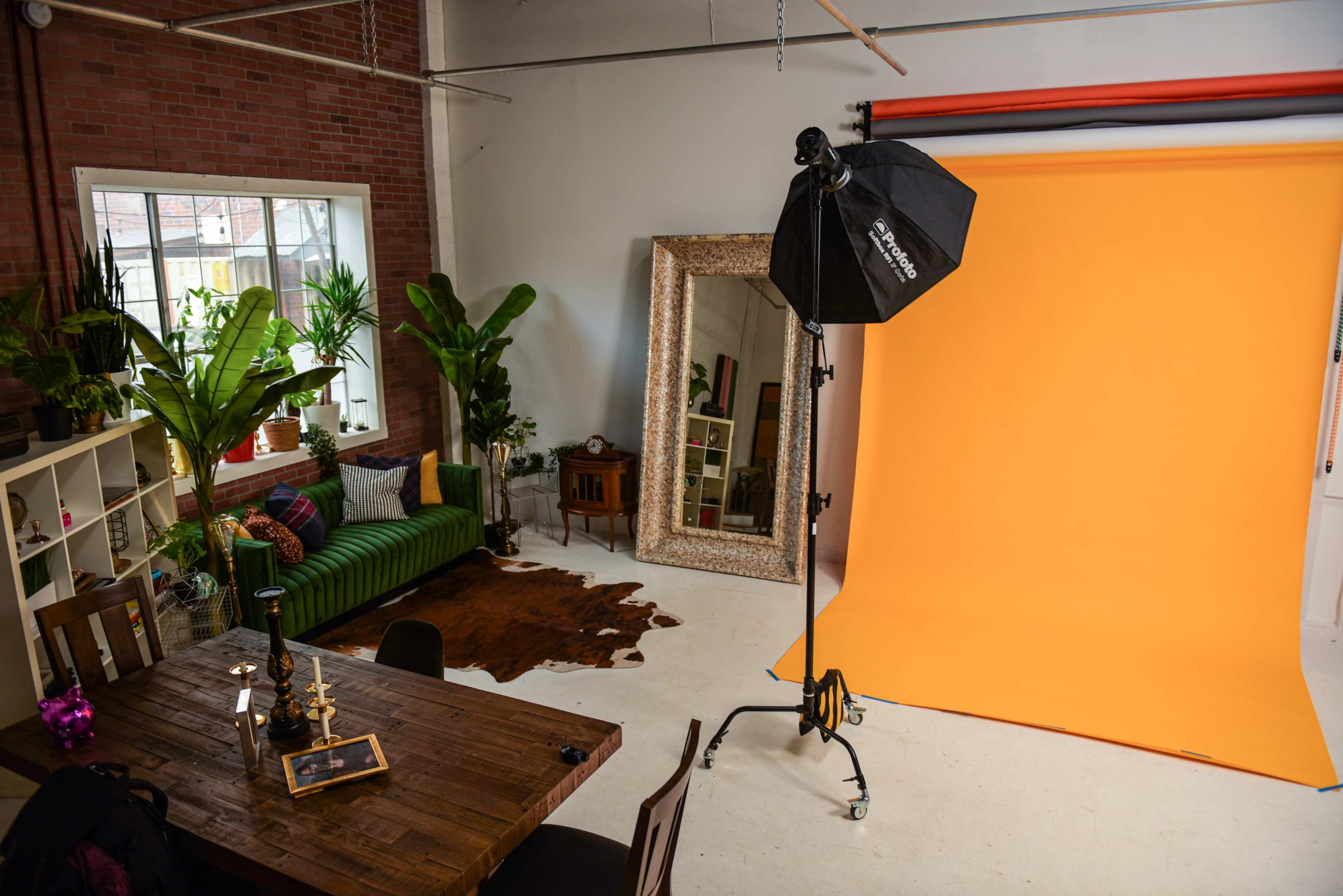 A photography studio featuring an orange backdrop, a green striped couch, a large mirror, tall plants, and a wooden table with decorative items.