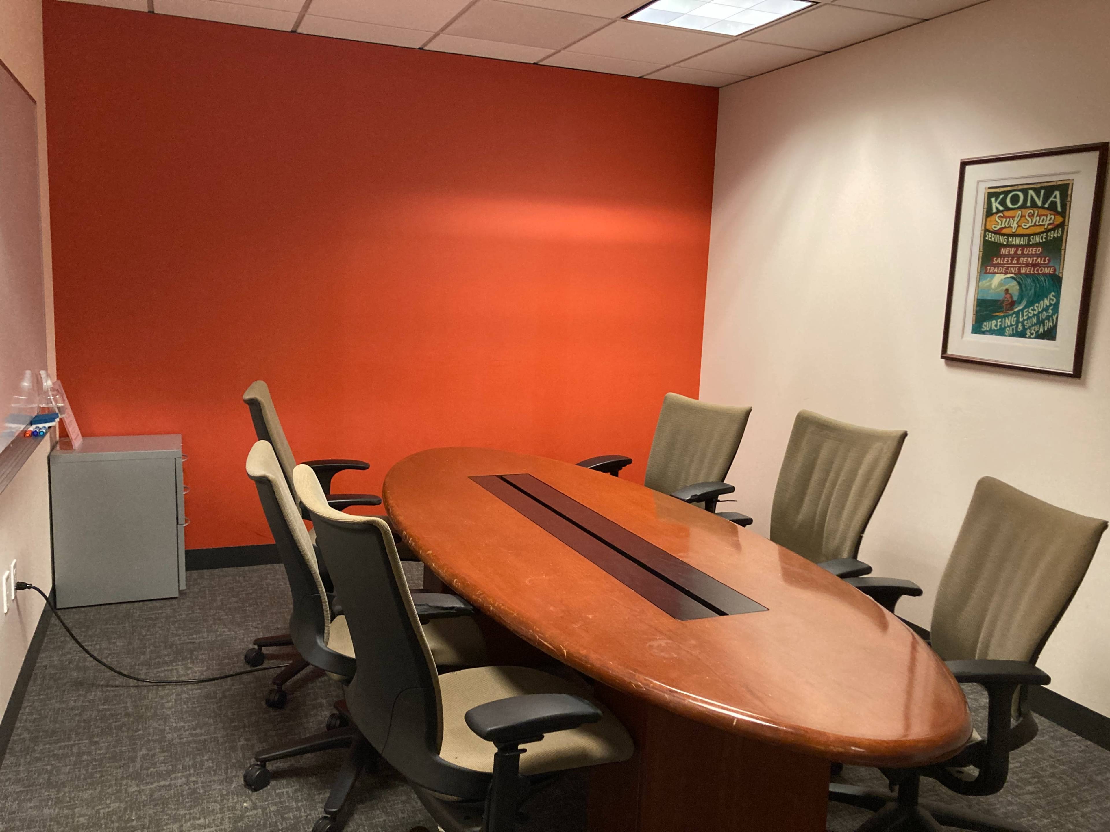 A conference room features a wooden oval table surrounded by six chairs and an orange accent wall.