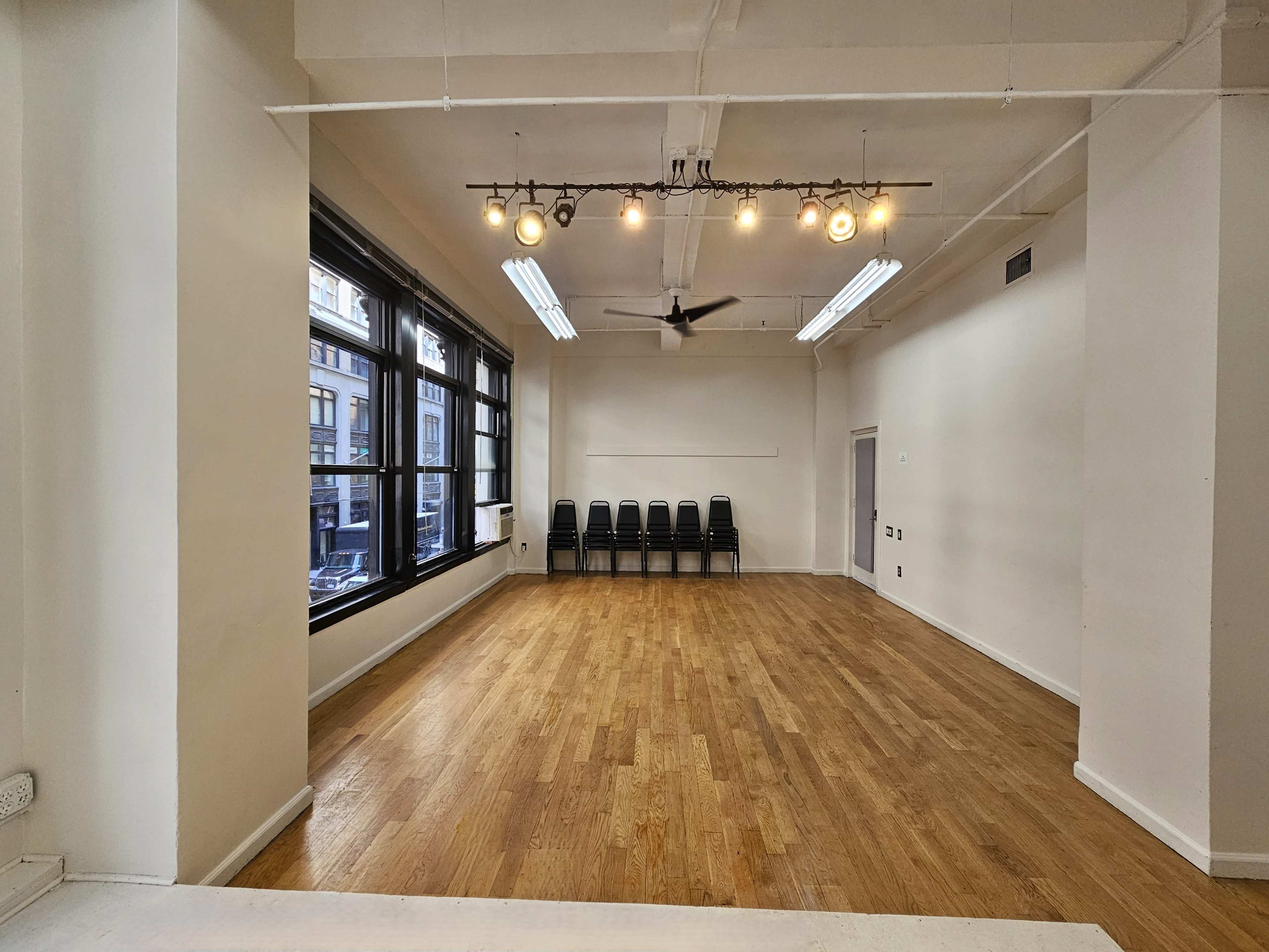An empty room with wooden flooring, large windows, a ceiling fan, and a row of black chairs against one wall.