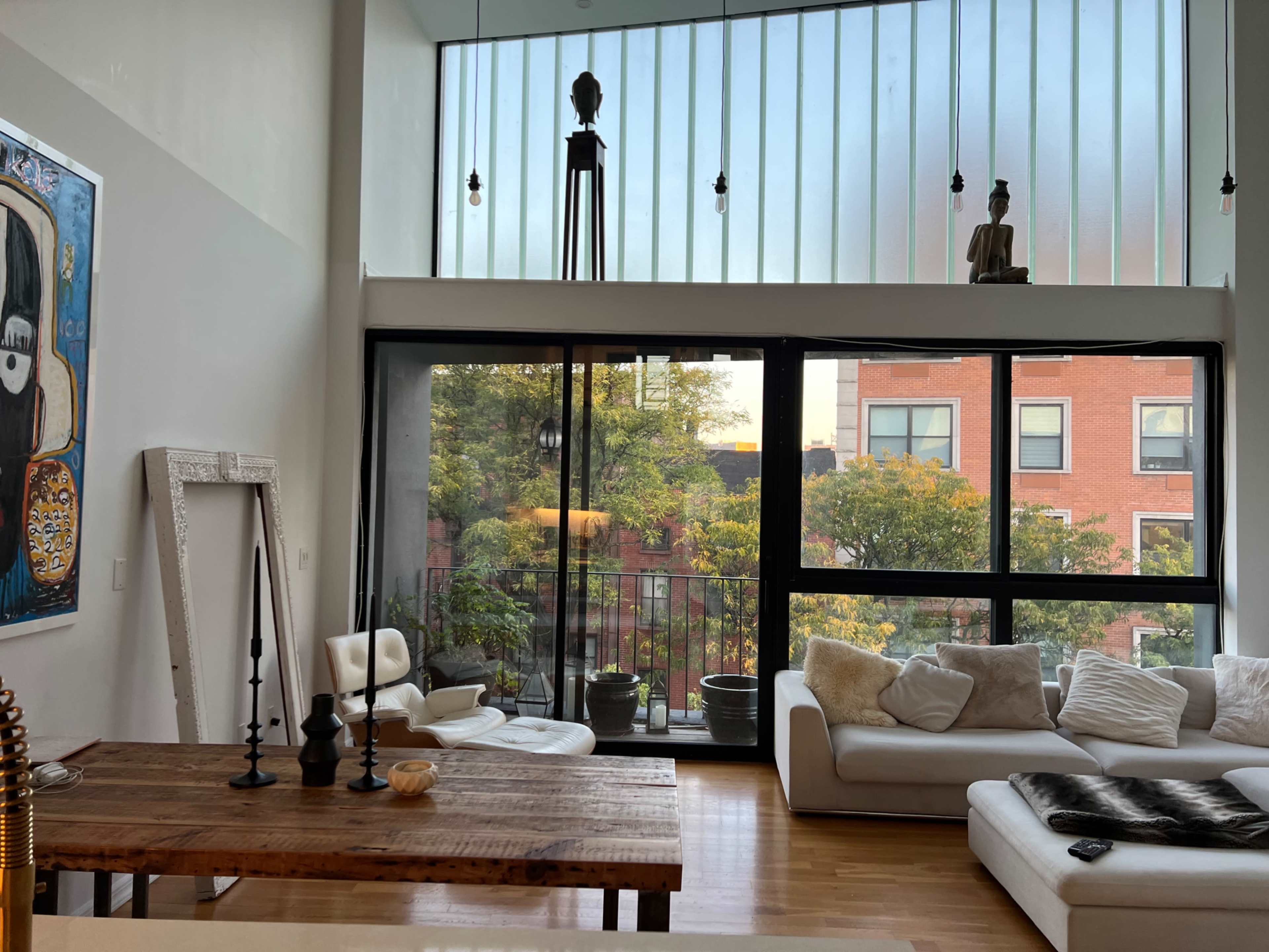 A stylish living room with large windows, a wooden dining table, white furniture, and potted plants.