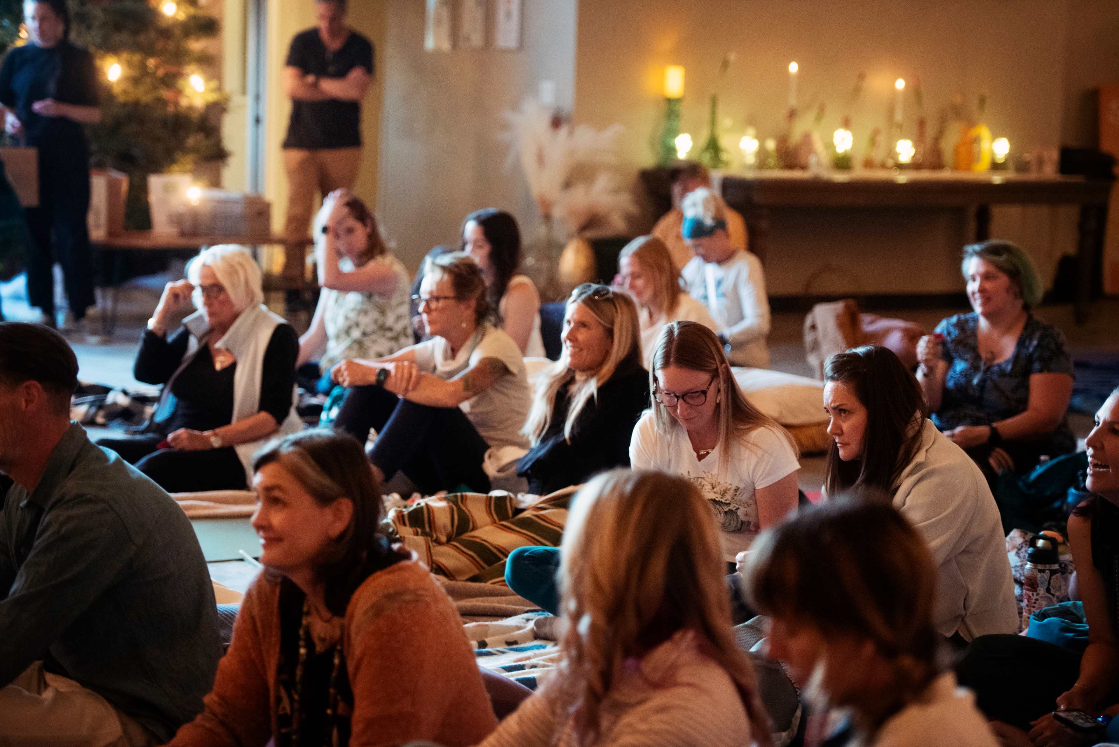 A diverse group of women and men sit attentively on the floor of a warmly lit room during a gathering or event.
