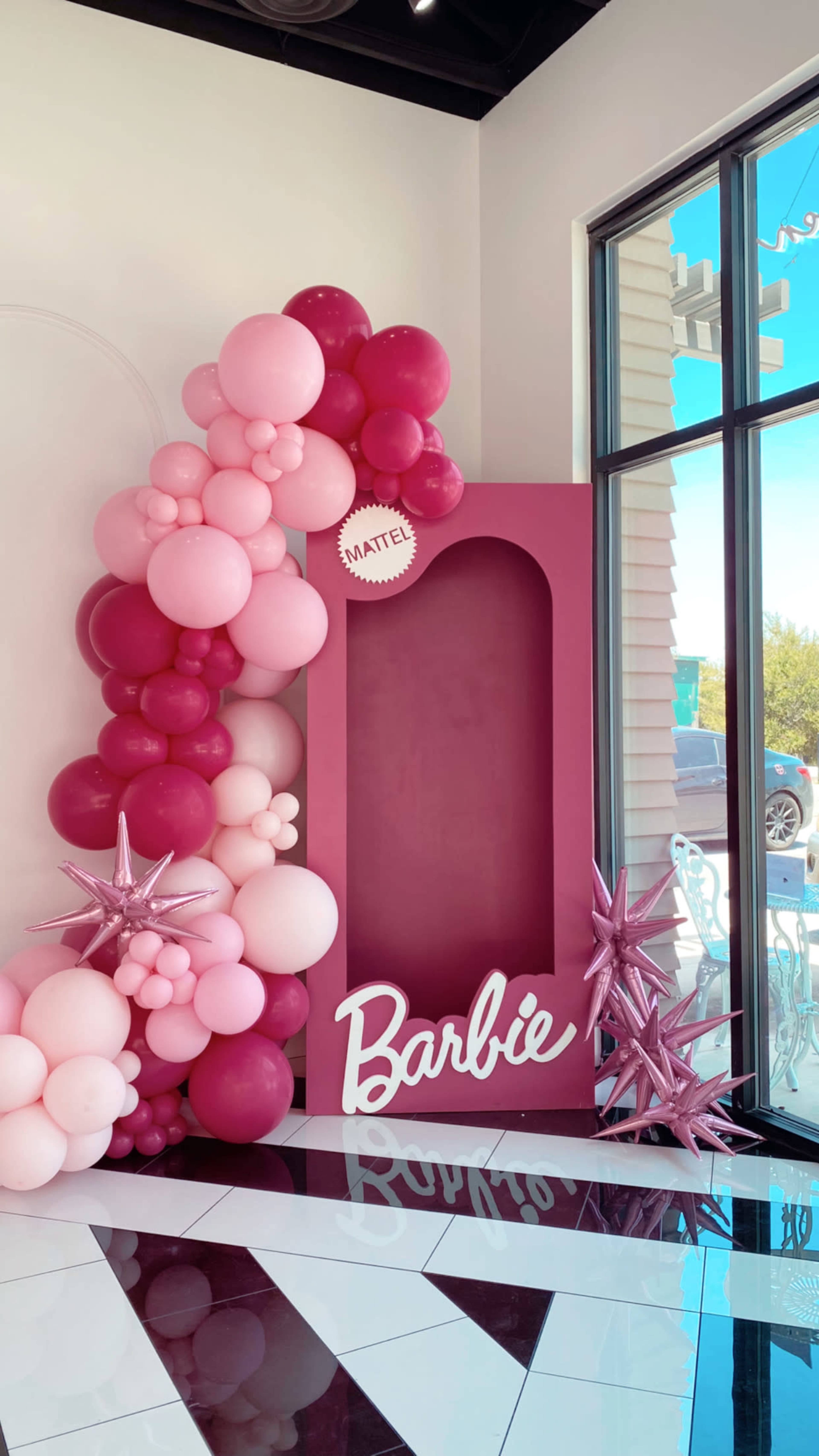 A decorative setup featuring a large pink and maroon balloon arch and a display frame with "Barbie" written on it, positioned next to a window.