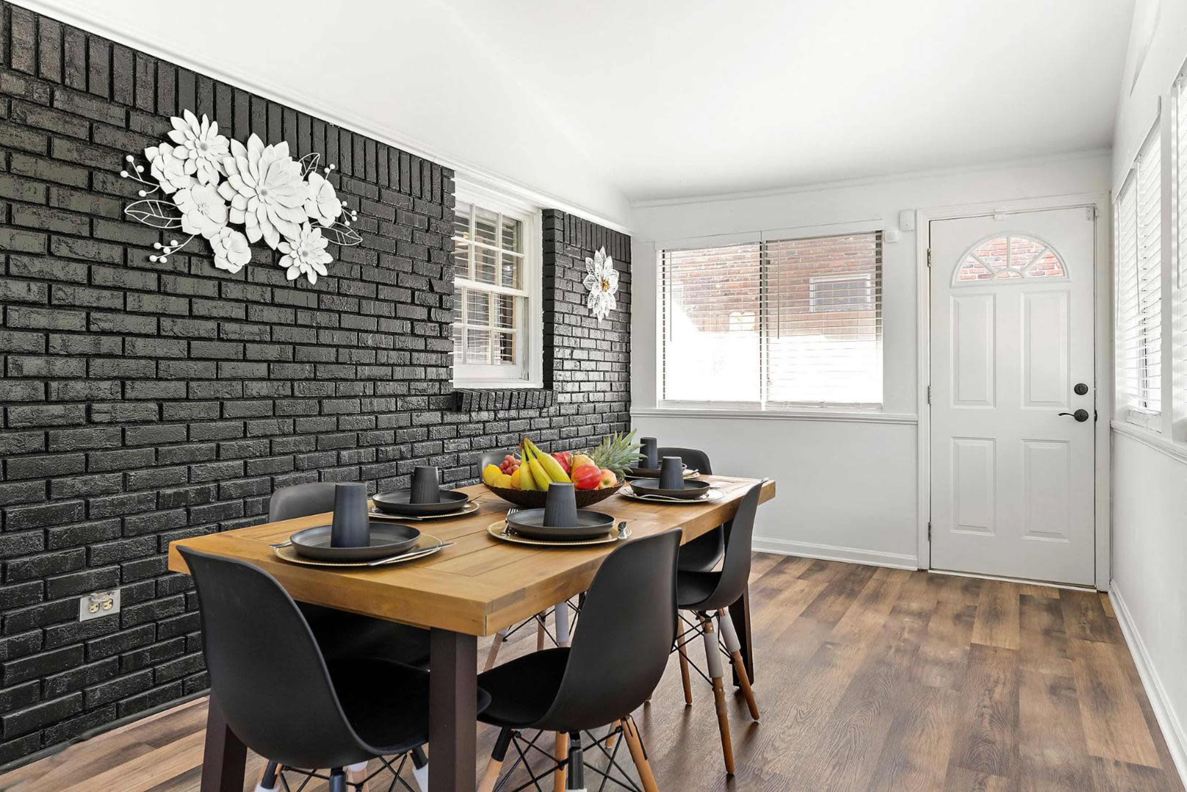 The image shows a dining area with a wooden table set for four, black chairs, and a decorative brick wall featuring white floral wall art.