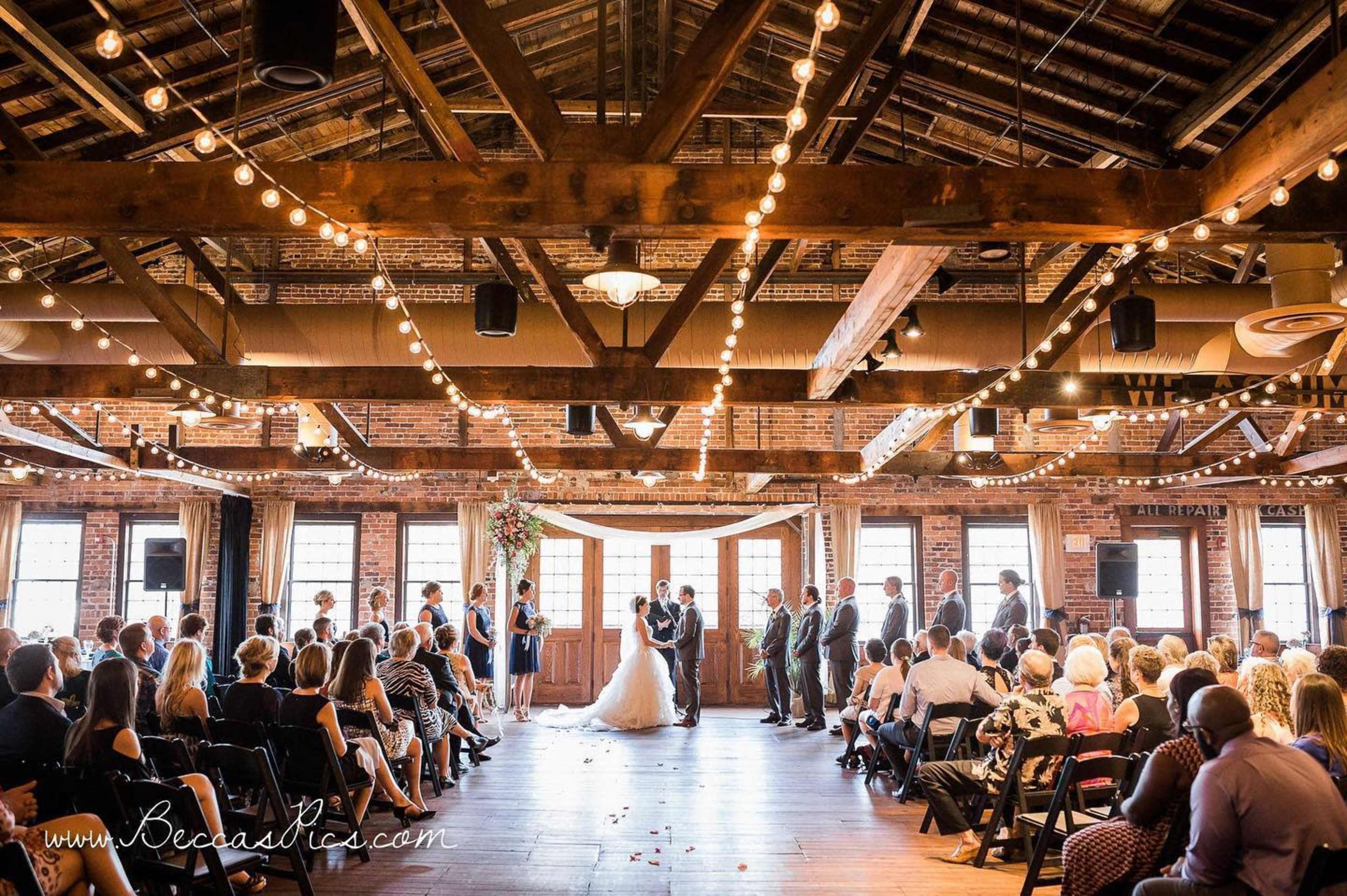A wedding ceremony takes place indoors in a decorated venue with exposed wooden beams and string lights, surrounded by seated guests.