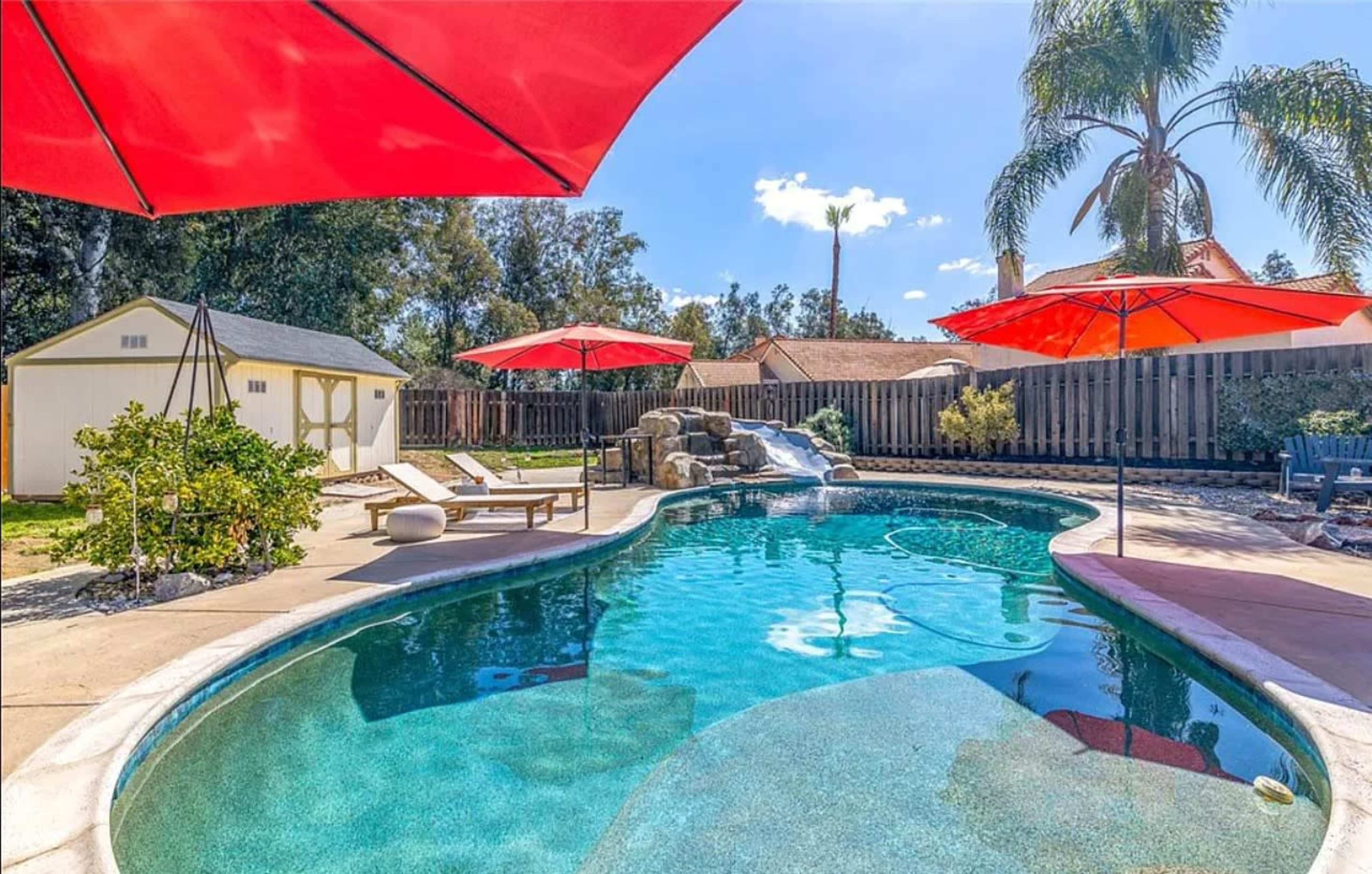 A backyard featuring a curved swimming pool with a waterslide, surrounded by red umbrellas and plants, alongside a shed in the background.