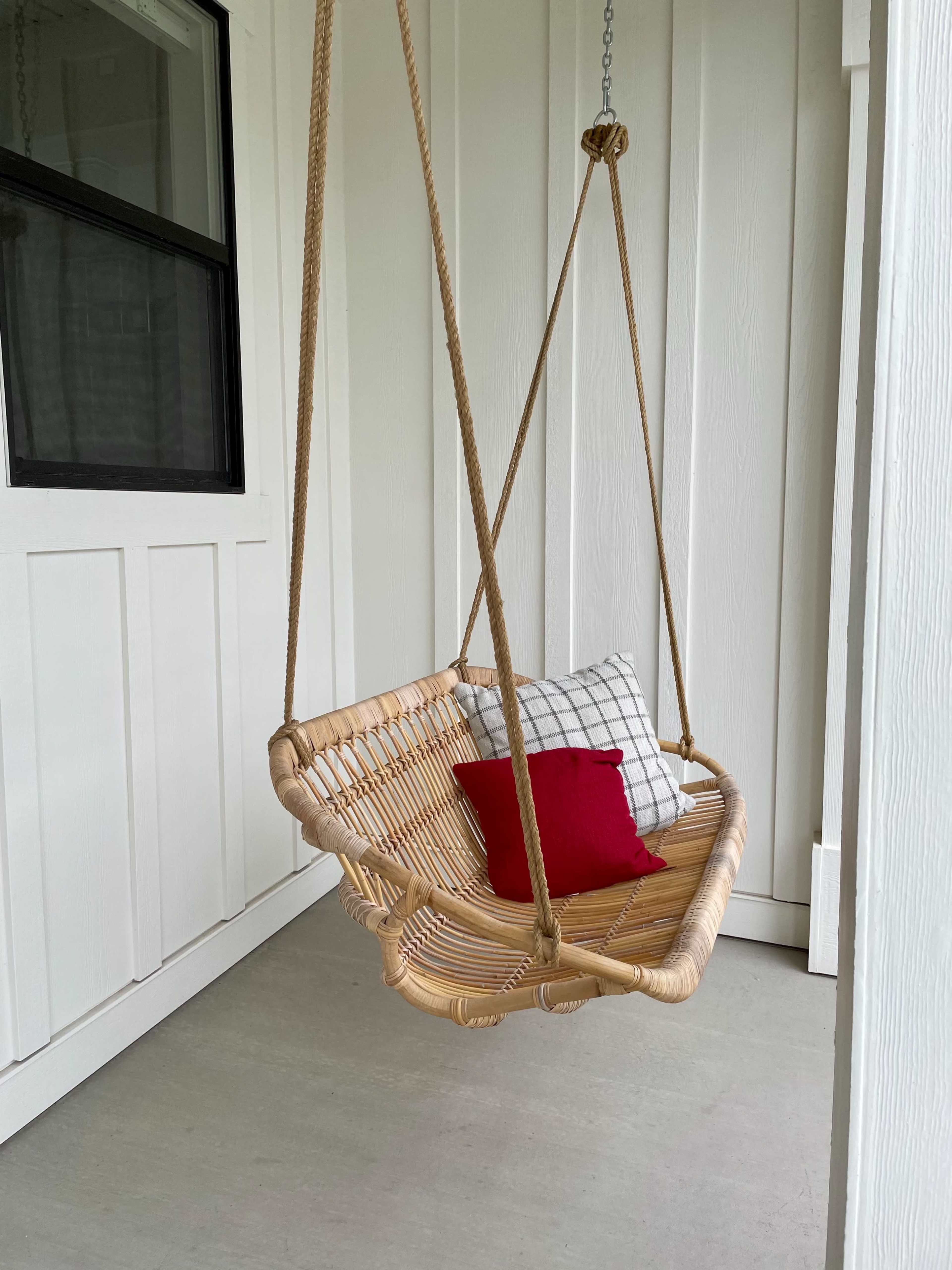 A woven hanging chair with a red and a checkered pillow is suspended from a ceiling in a white-paneled porch.