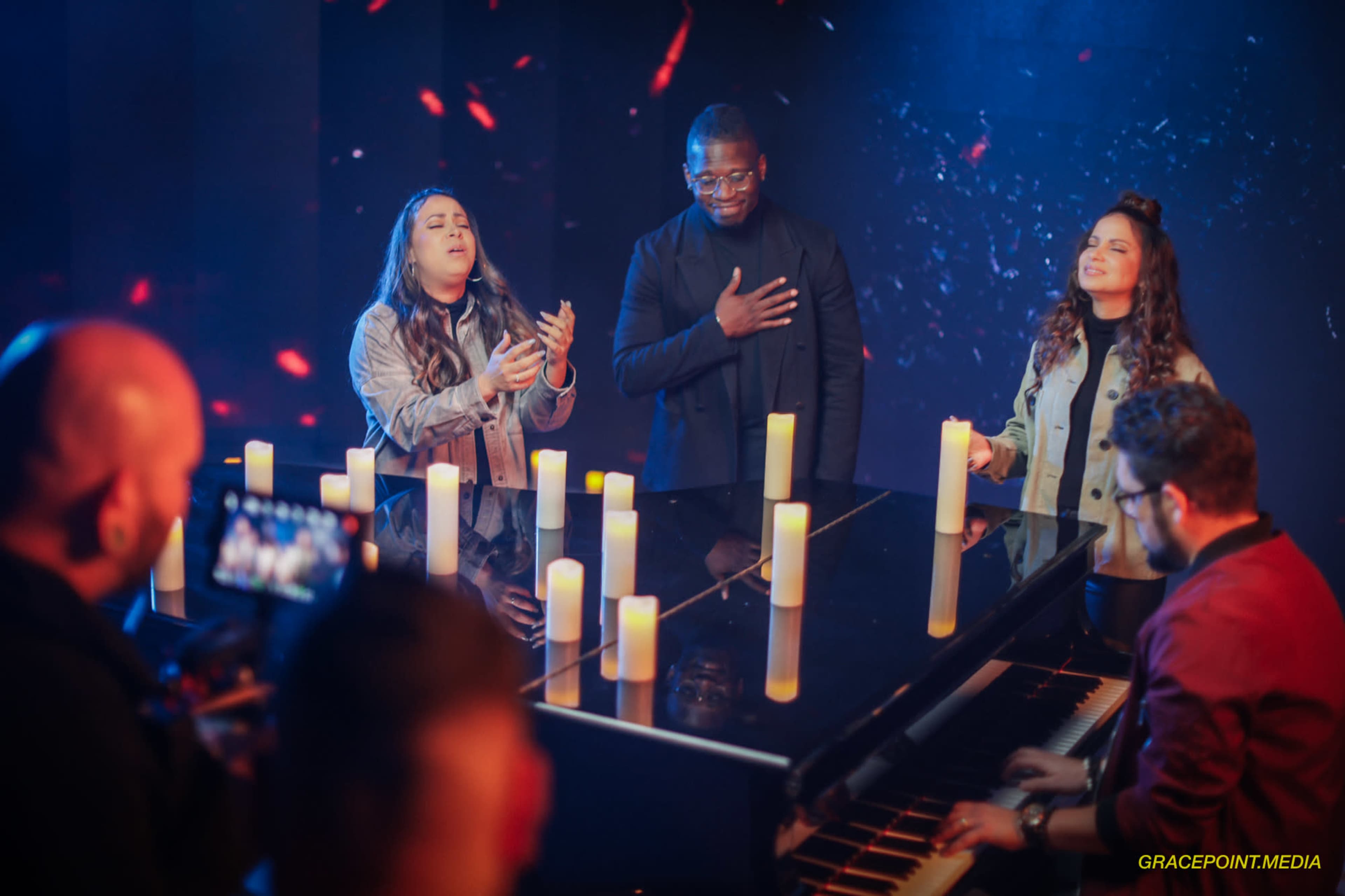 A group of musicians performs around a piano with candles lit in front of them, set against a dark background with colorful lights.