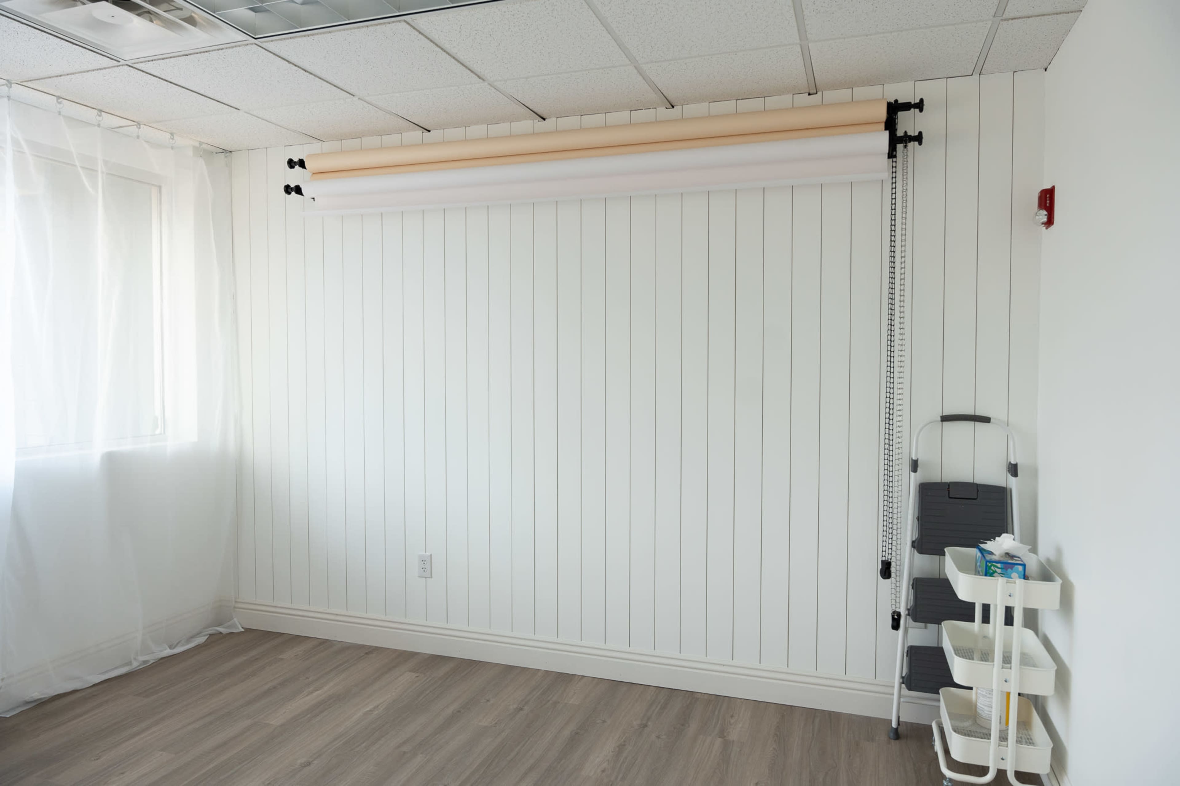 The image shows a plain, well-lit room with a white paneled wall, a backdrop support system with three rolls of fabric, and a small cart with supplies.