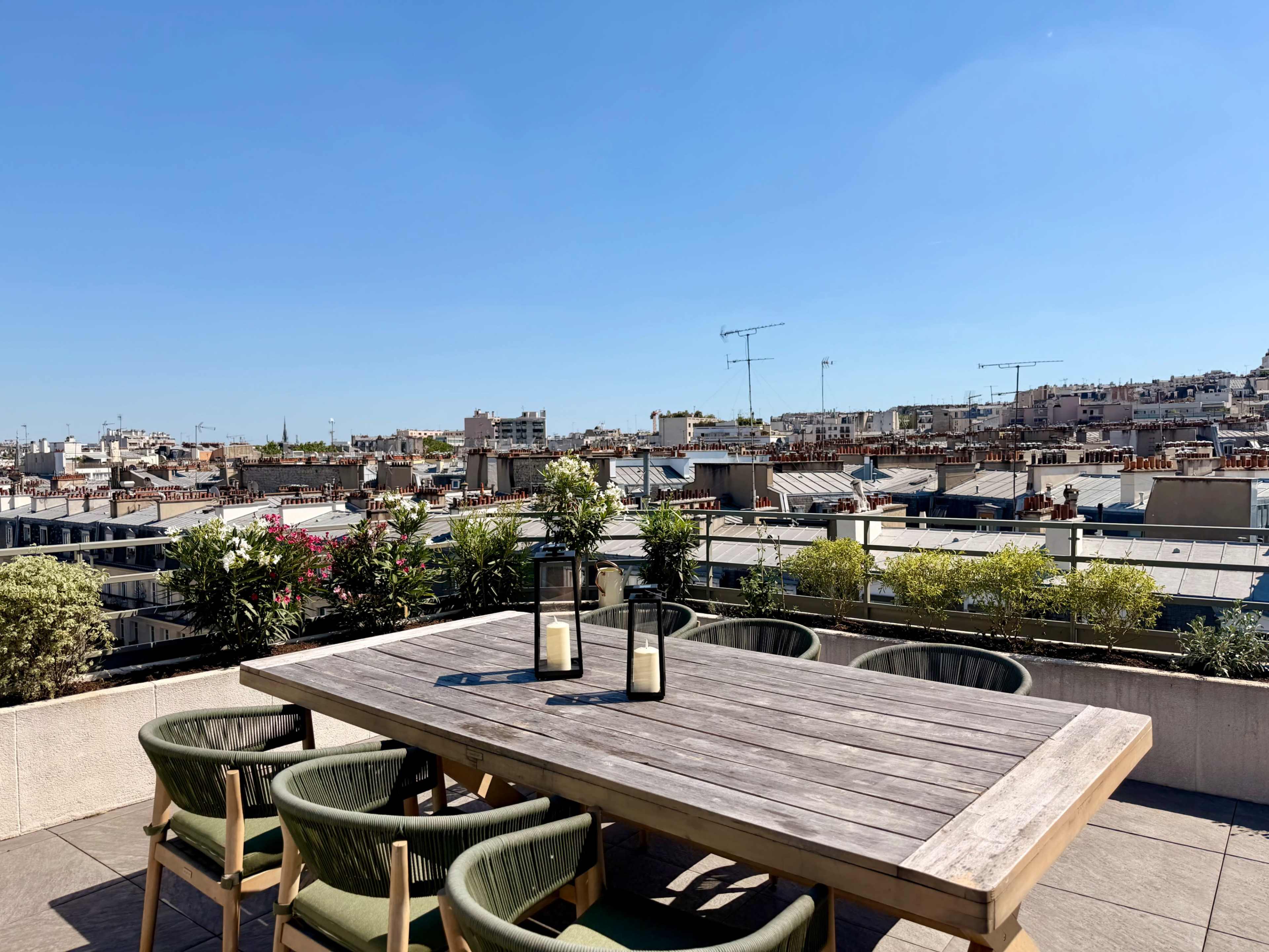 A rooftop terrace features a wooden dining table surrounded by green chairs, with a panoramic view of city rooftops and a clear blue sky.
