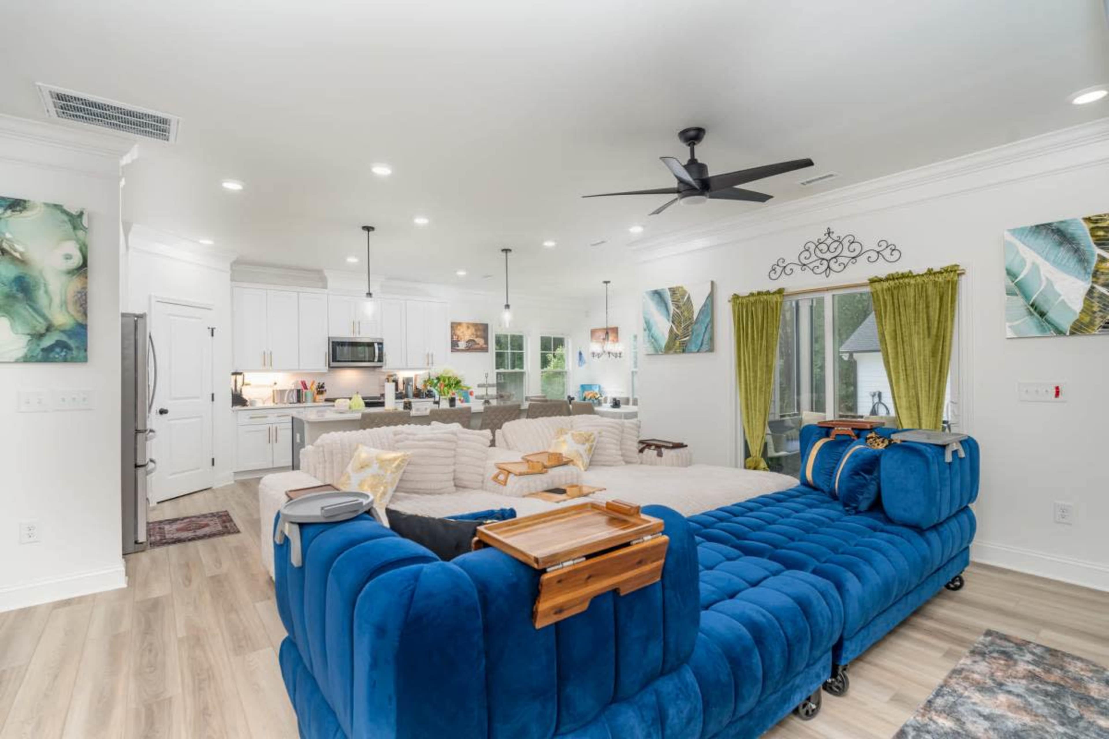 The image shows a modern living room with a blue sectional sofa, a large open space that connects to a kitchen, and bright green curtains.