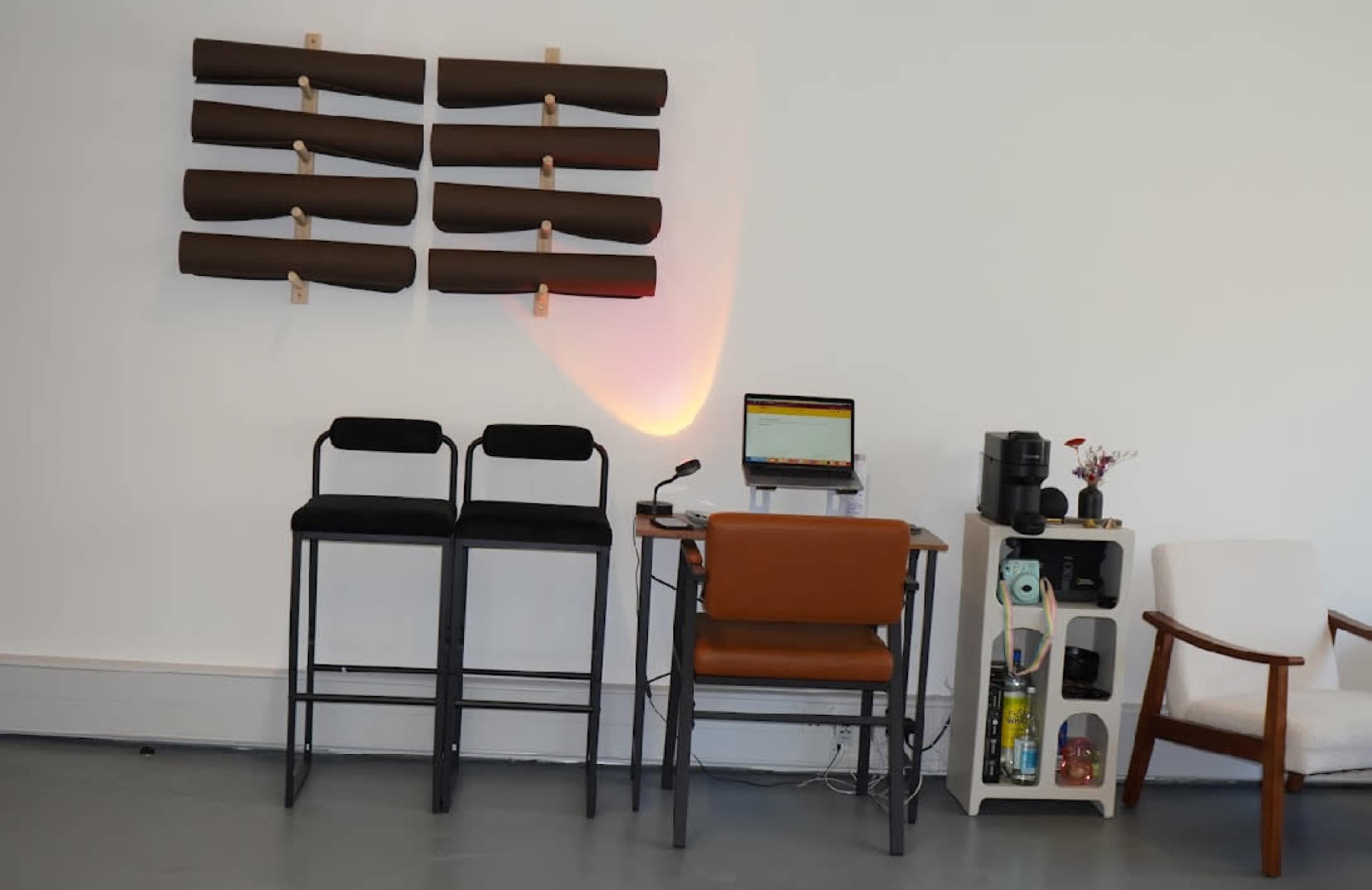 The image shows a modern workspace with two black stools, a brown desk chair, a desk with a computer and microphone, and a wall-mounted display of rolled brown papers.