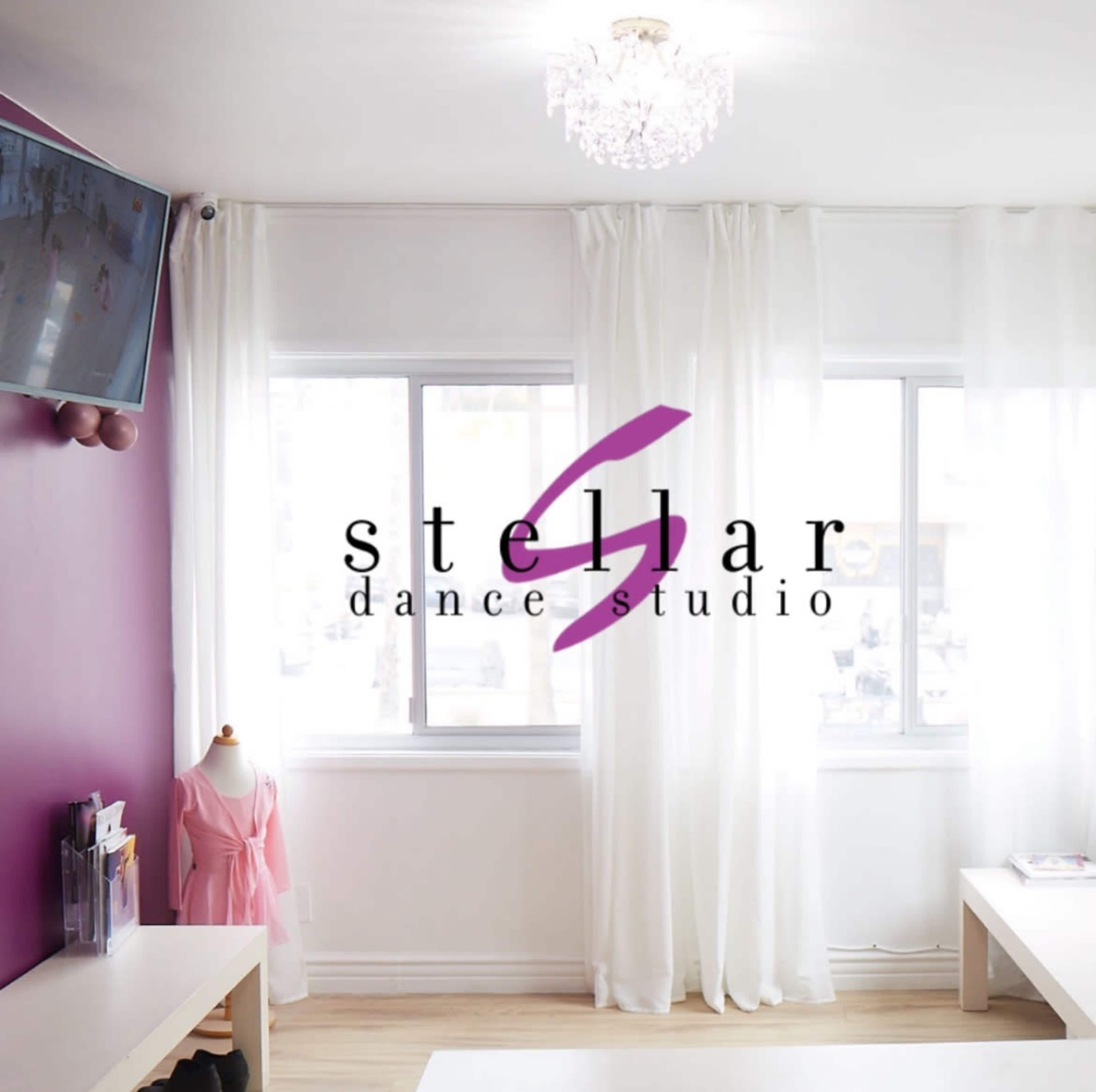 A dance studio with a purple accent wall, large windows covered by white curtains, and a small dress form displaying a pink garment.