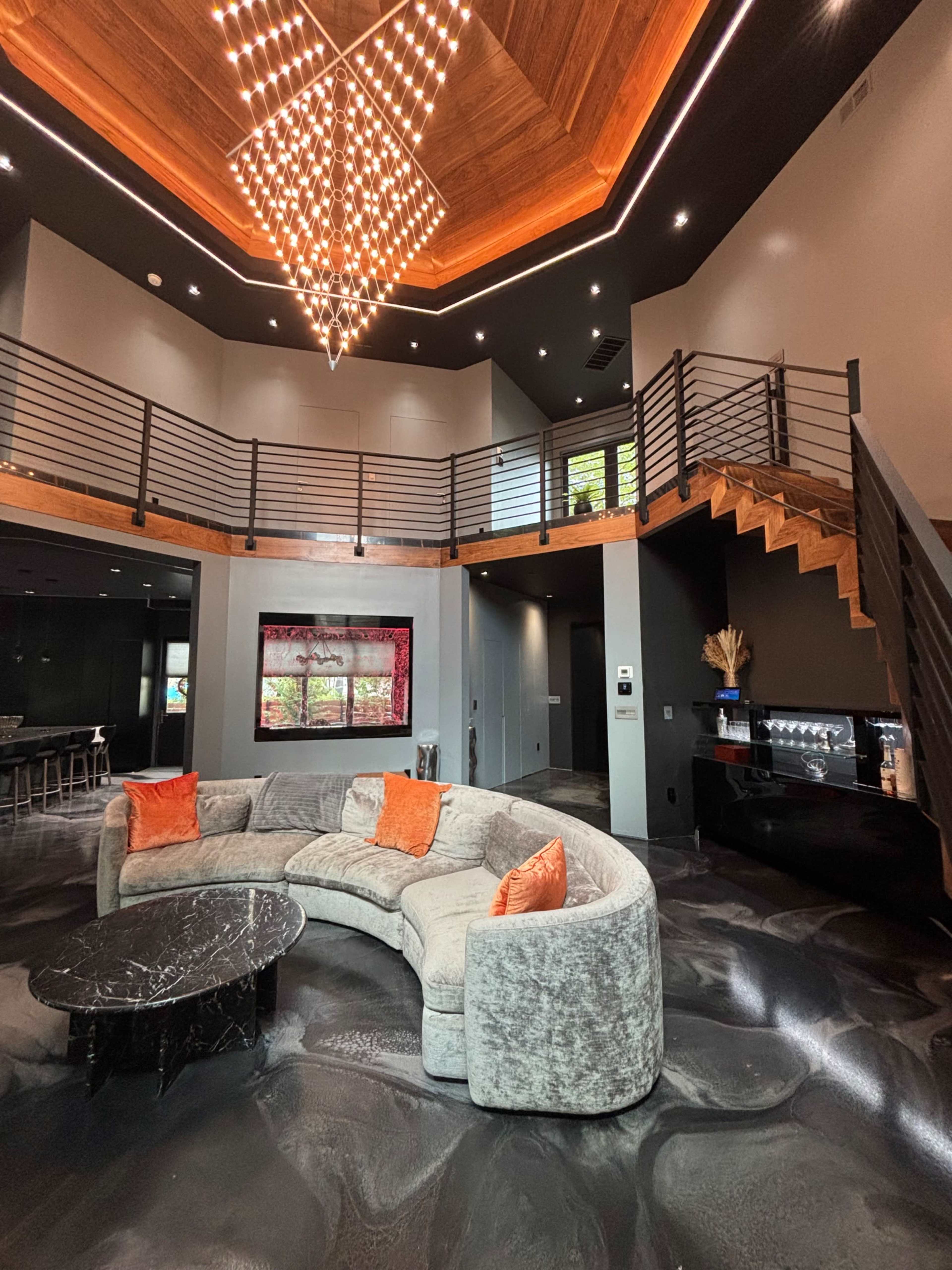 The image shows a modern interior living area featuring a curved sofa, a black marble coffee table, a striking chandelier, and a staircase leading to an upper level.