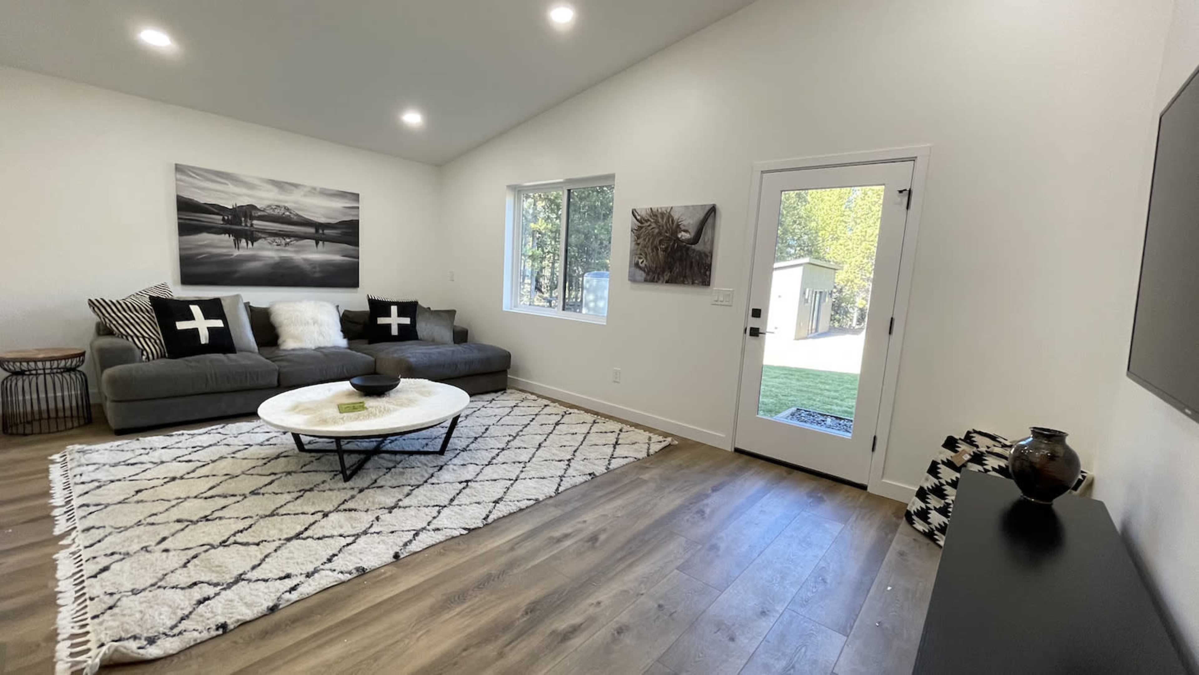 A modern living room with a gray sectional sofa, a round coffee table on a patterned rug, and a glass door leading to an outdoor area.