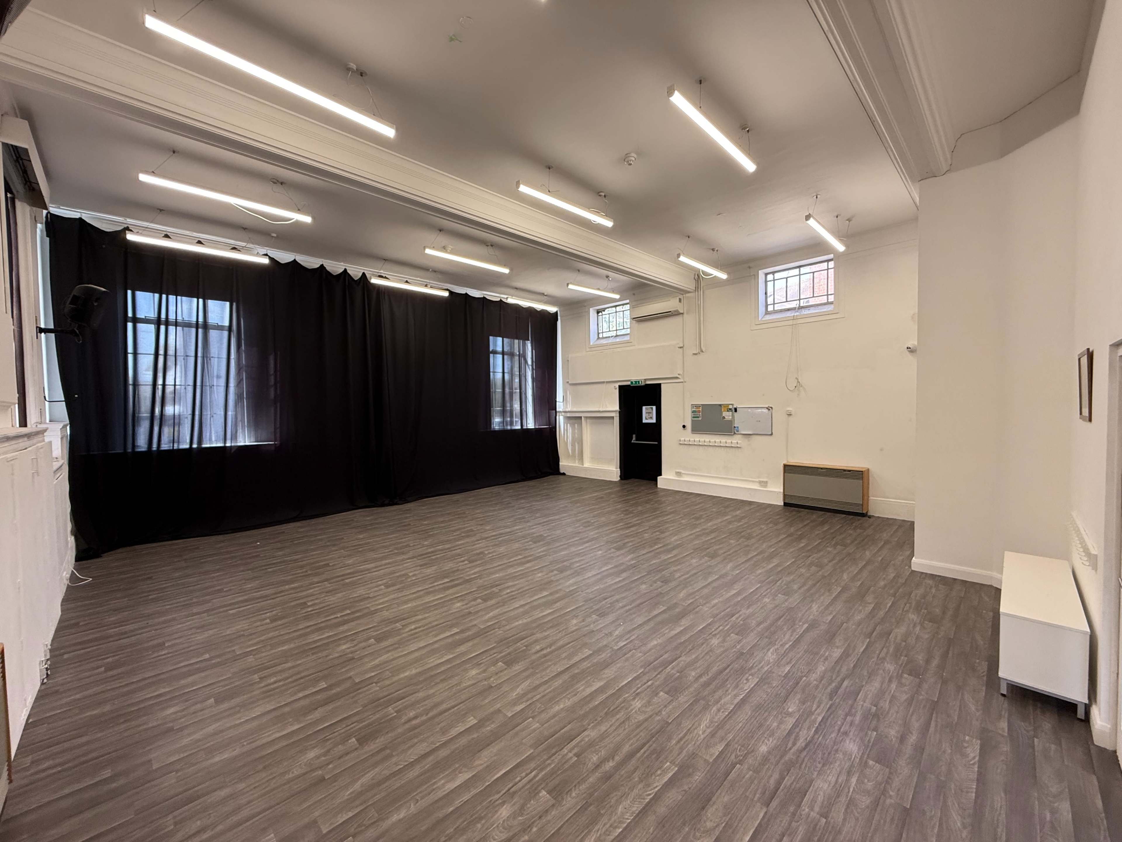 The image shows a spacious, empty room with gray wooden flooring, white walls, and large windows covered by black curtains.