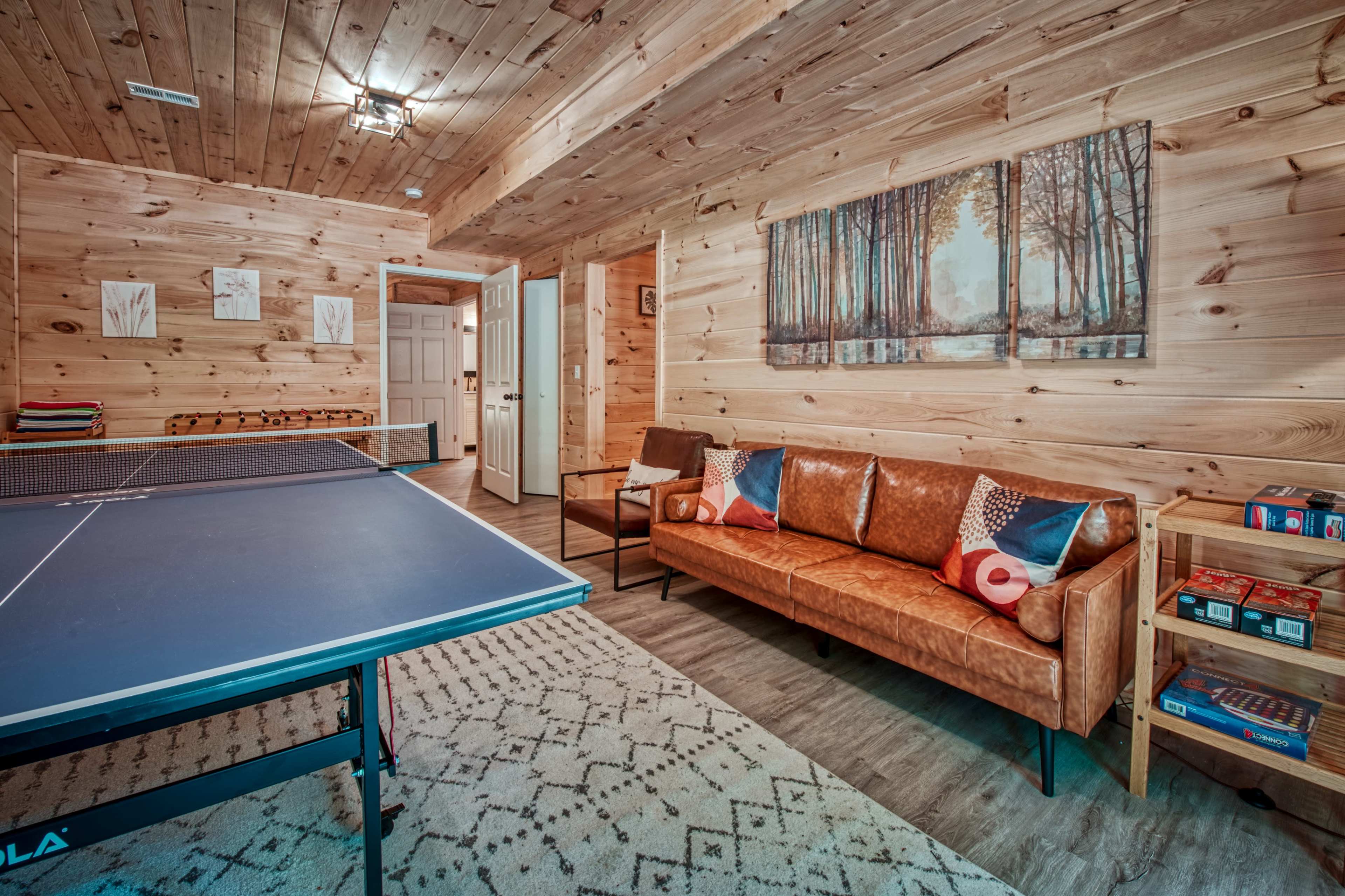 A cozy game room featuring a ping pong table, a leather sofa, and wooden wall paneling, with a painting of a forest scene on the wall.