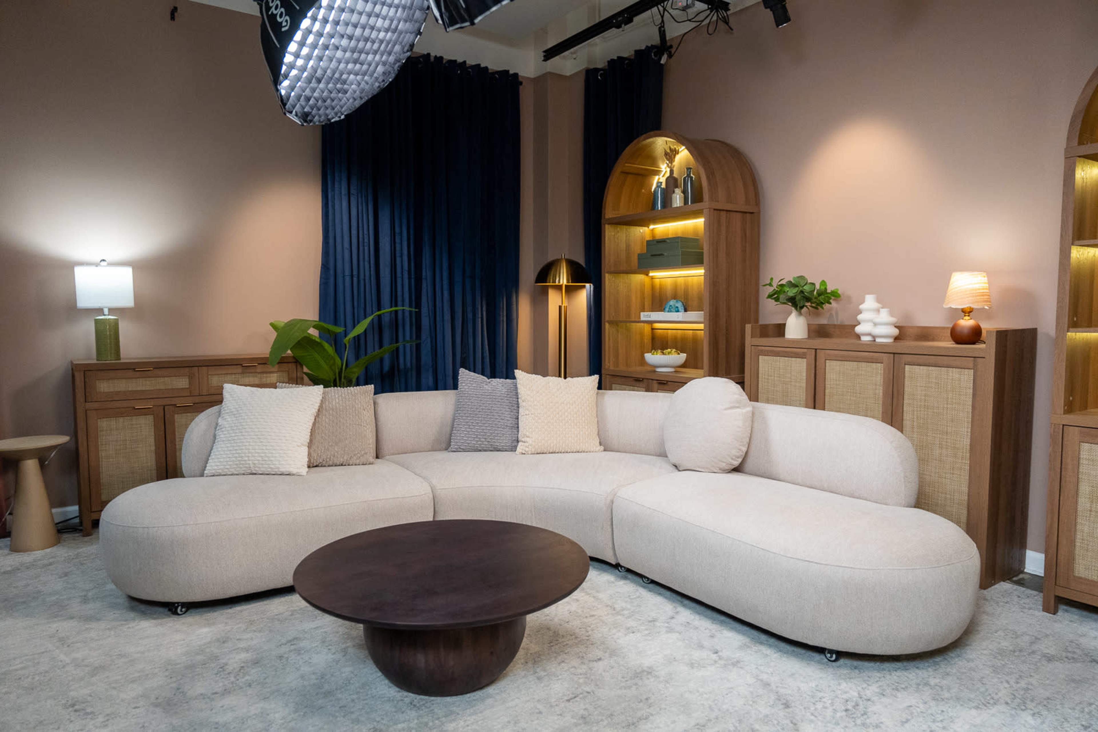 The image shows a modern living room with a curved beige sofa, a round wooden coffee table, and wooden shelving with decorative items against a backdrop of blue curtains.