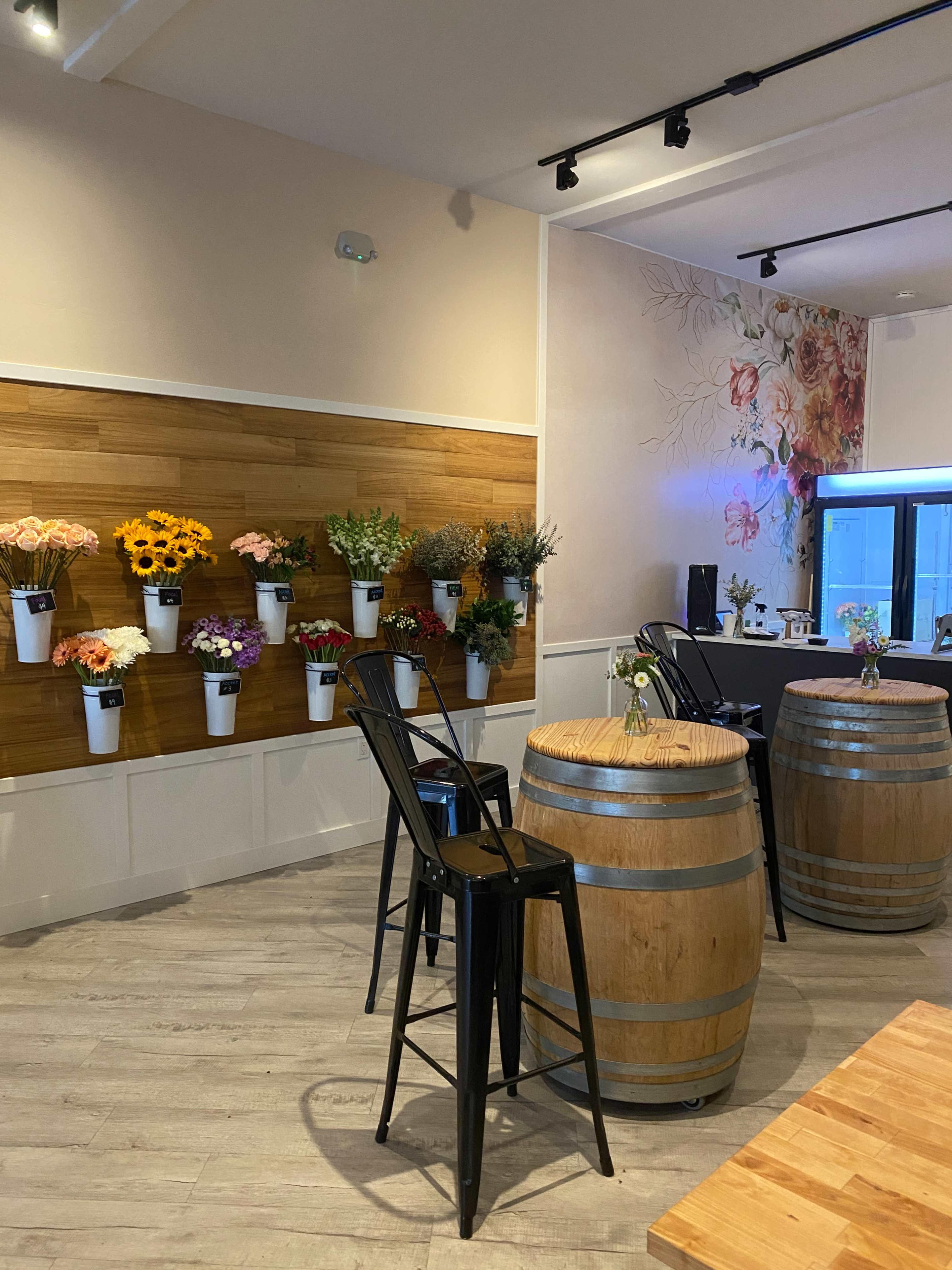 The image shows a cozy café interior featuring flower arrangements displayed on a wooden wall and wooden barrels serving as tables.