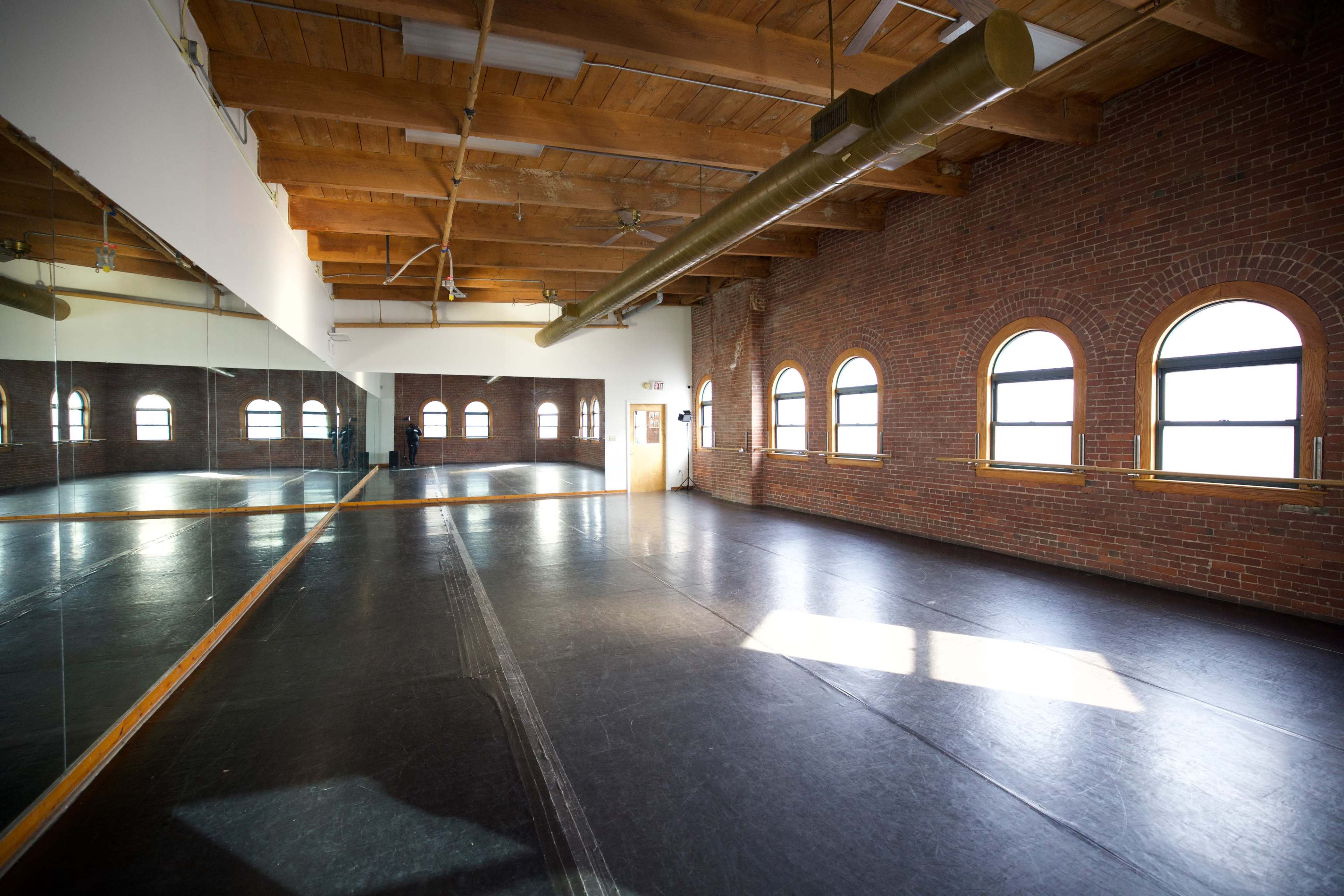 A spacious dance studio with wooden beams, a mirrored wall, and arched windows letting in natural light.