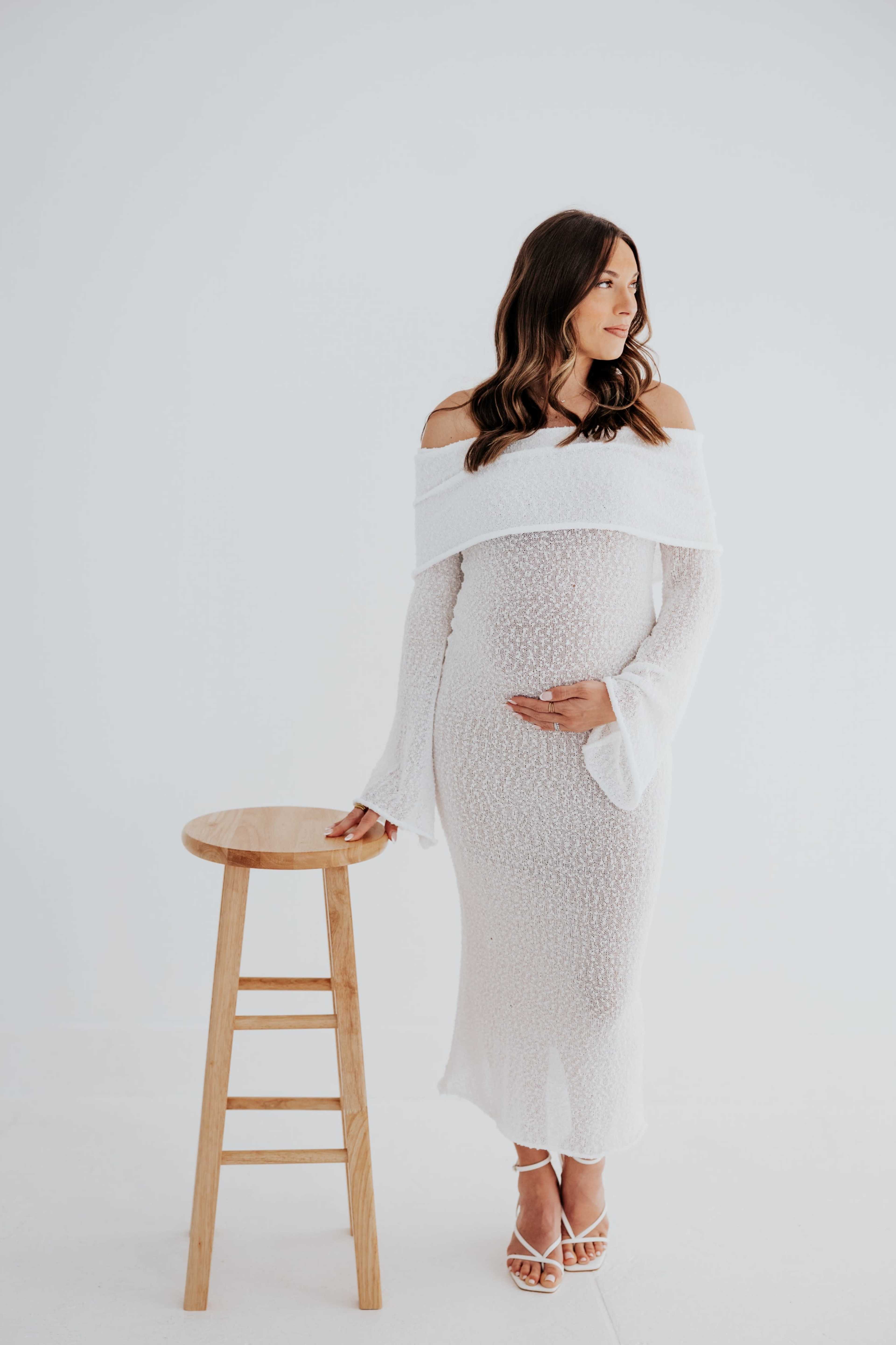 A woman in a white off-the-shoulder dress stands next to a wooden stool, holding her belly with one hand.