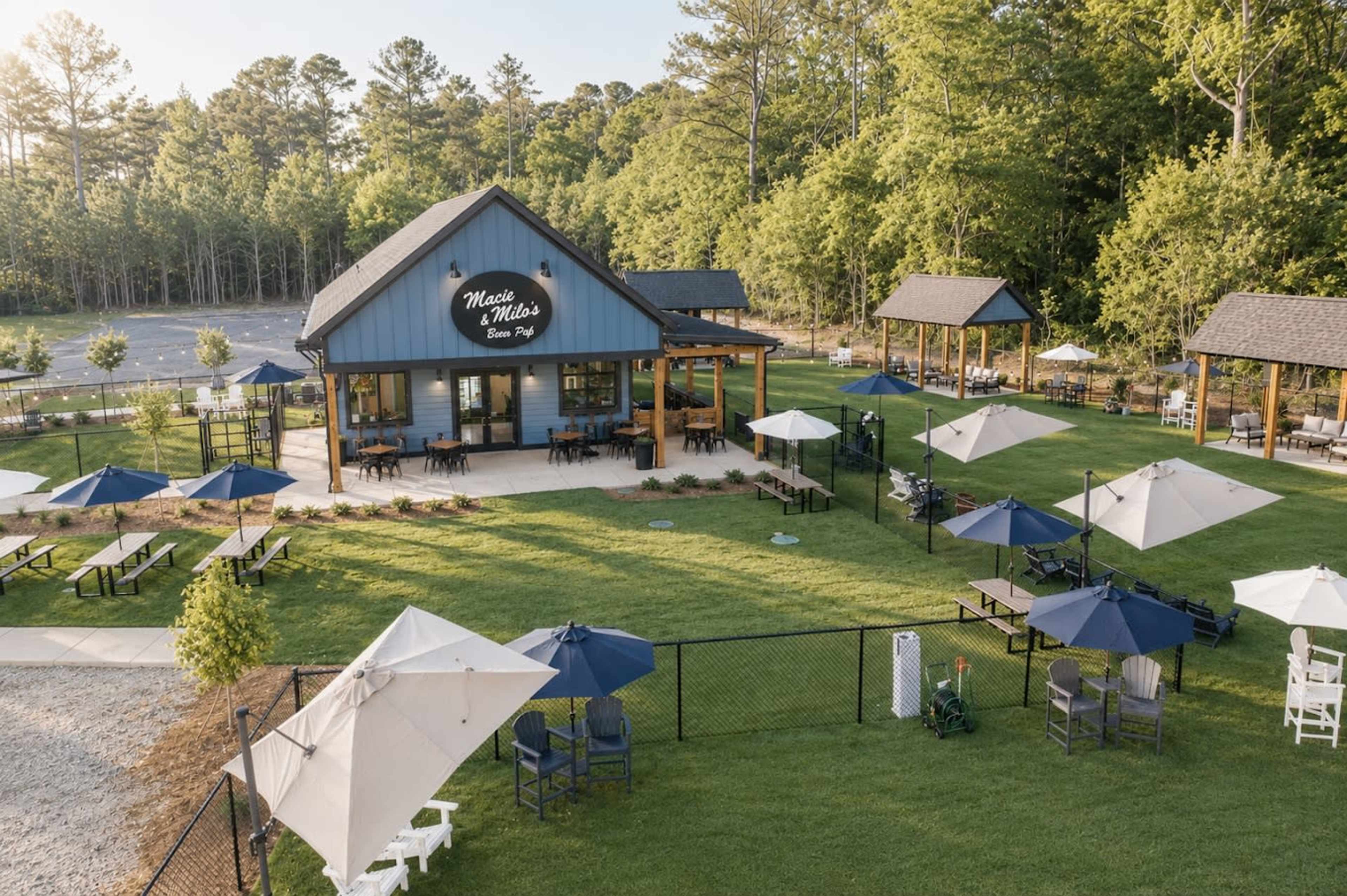 The image shows a blue barn-style building with outdoor seating, umbrellas, and landscaping in a green area surrounded by trees.