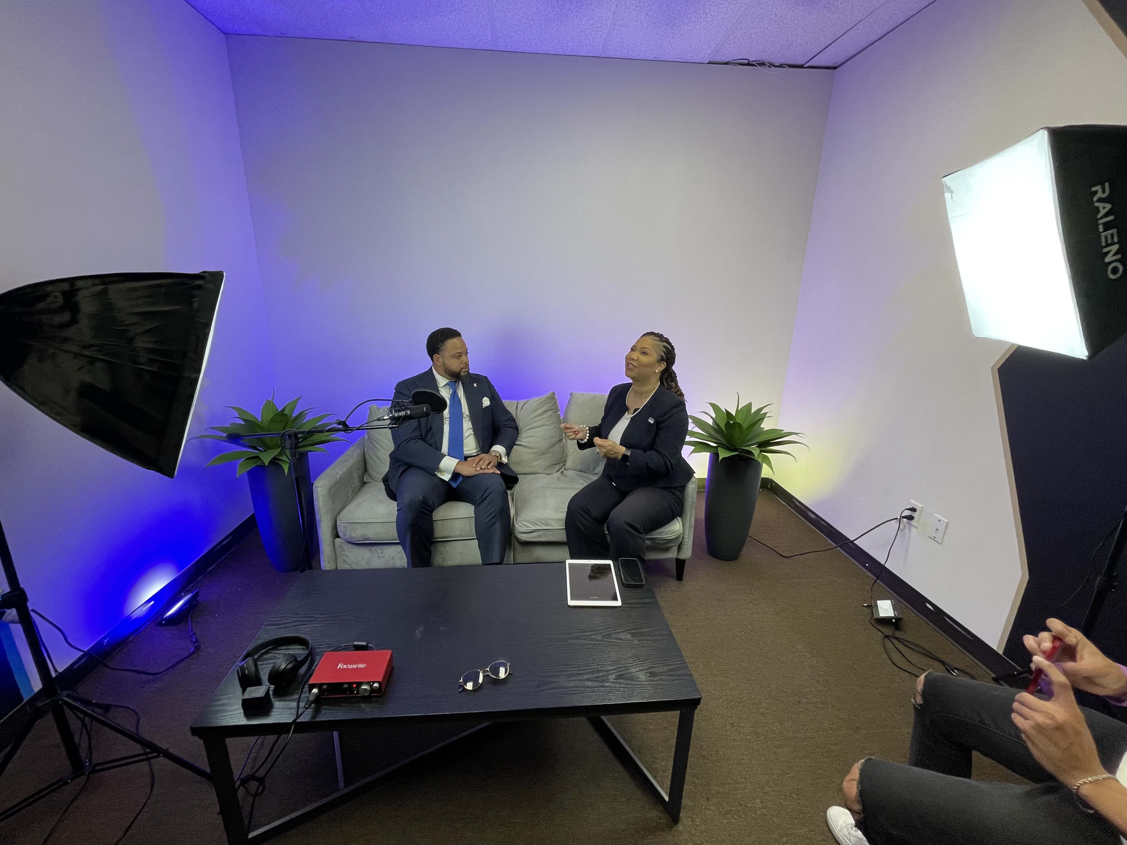 A man and a woman sit on a couch in a studio setting, engaged in discussion under professional lighting with potted plants in the background.