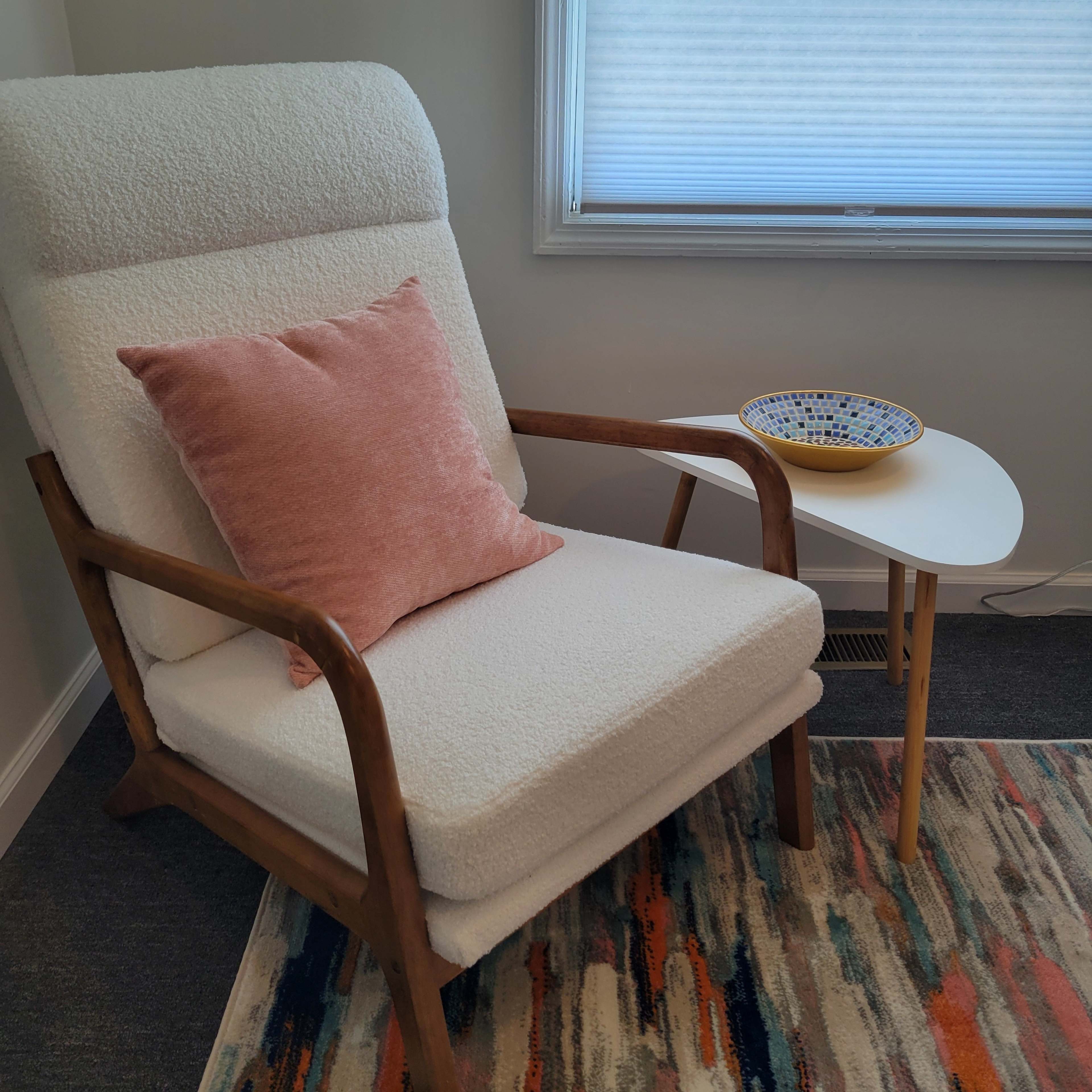 A white upholstered chair with a pink pillow sits beside a small round table holding a patterned bowl, on a colorful rug in a well-lit room.