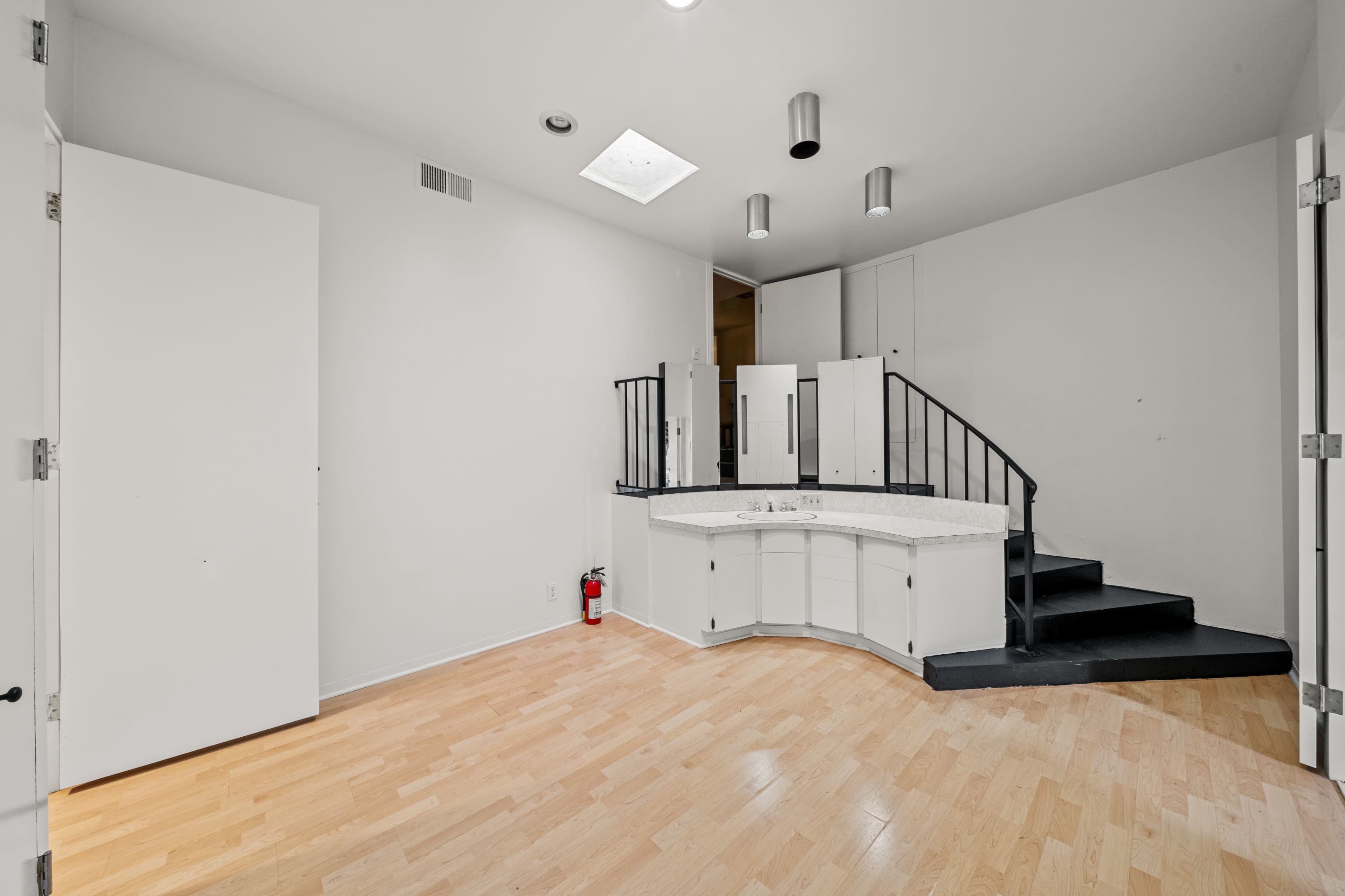 The image shows an empty room with light-colored wooden flooring, a curved staircase leading to a higher level, and a small bar area with a countertop.