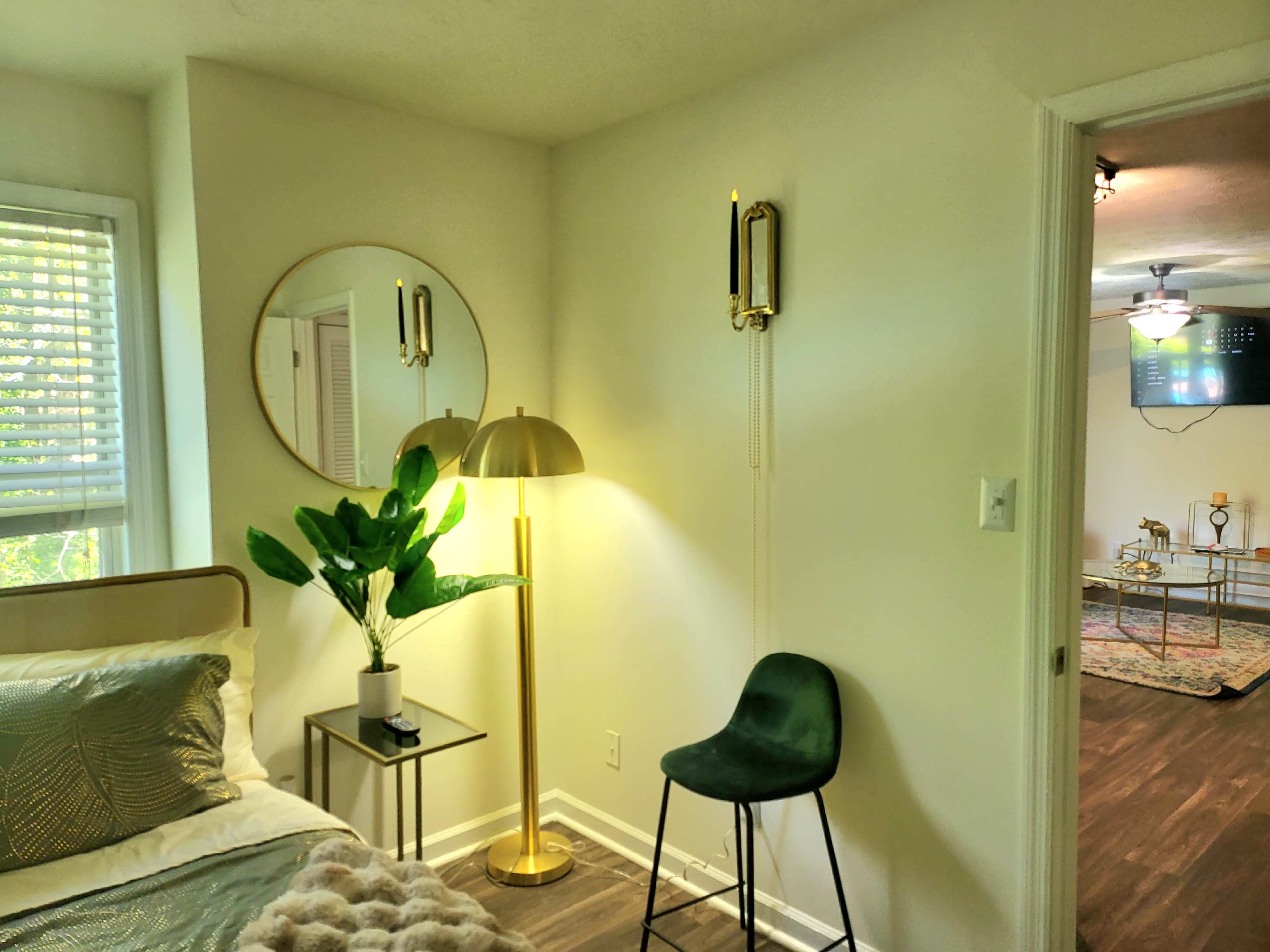A well-lit bedroom featuring a bed, a round mirror on the wall, a floor lamp, a plant, and a green chair, with a glimpse of a living room in the background.