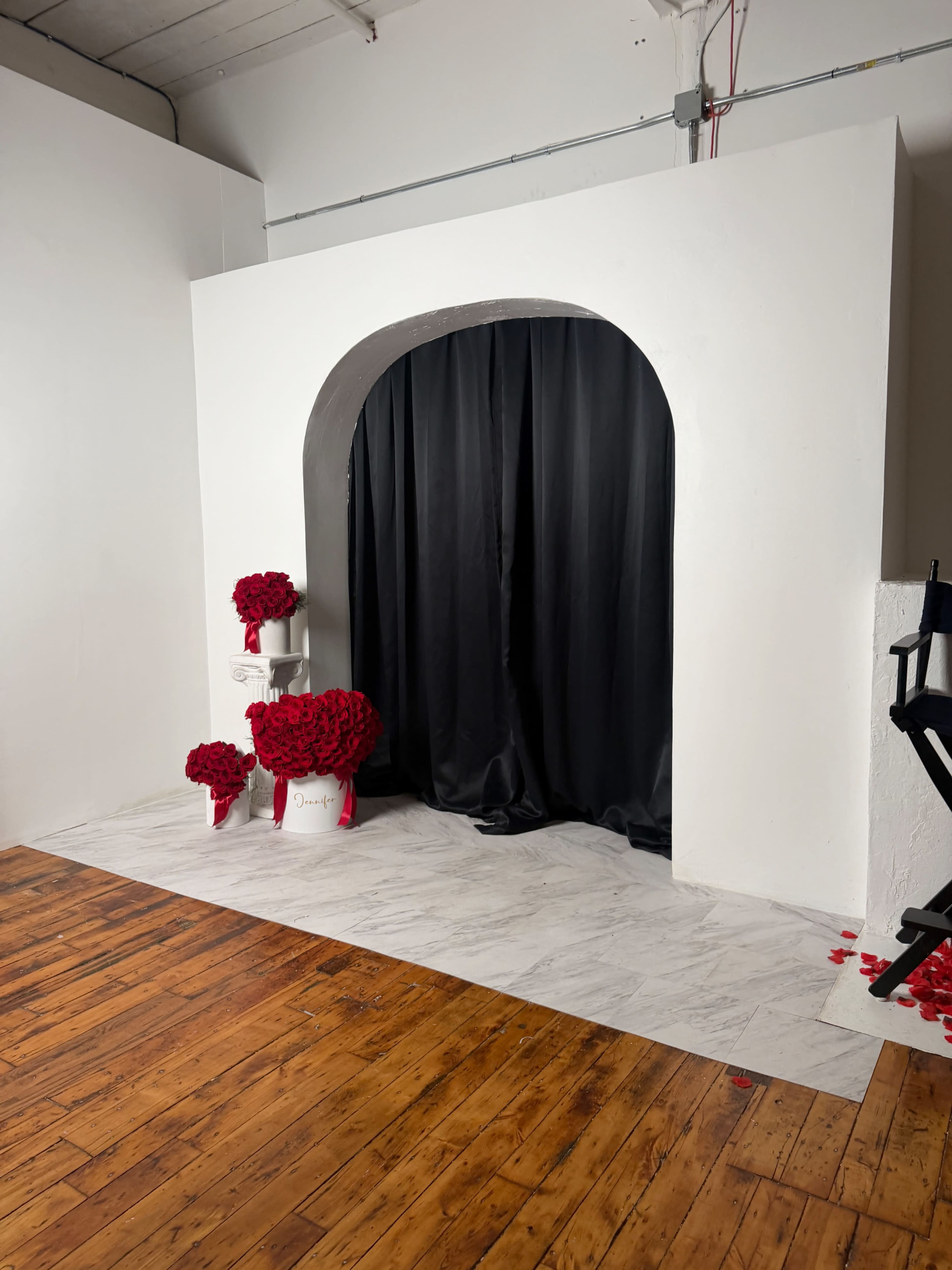 The image shows a white-walled space featuring a black curtain entrance with red flower arrangements positioned nearby on a wooden floor.