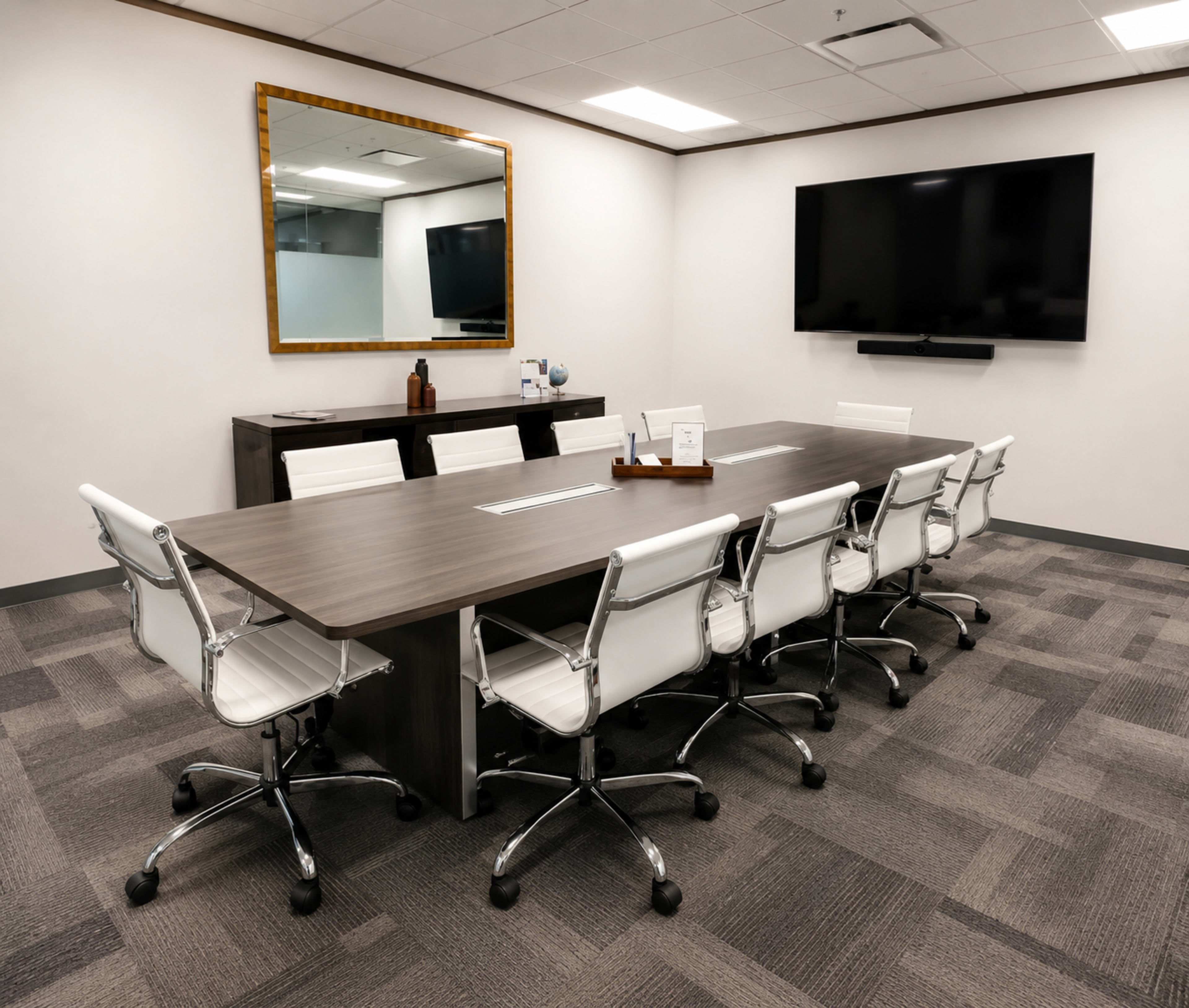 The image shows a modern conference room with a large rectangular table surrounded by white ergonomic chairs, a wall-mounted television, and a mirror reflecting the space.