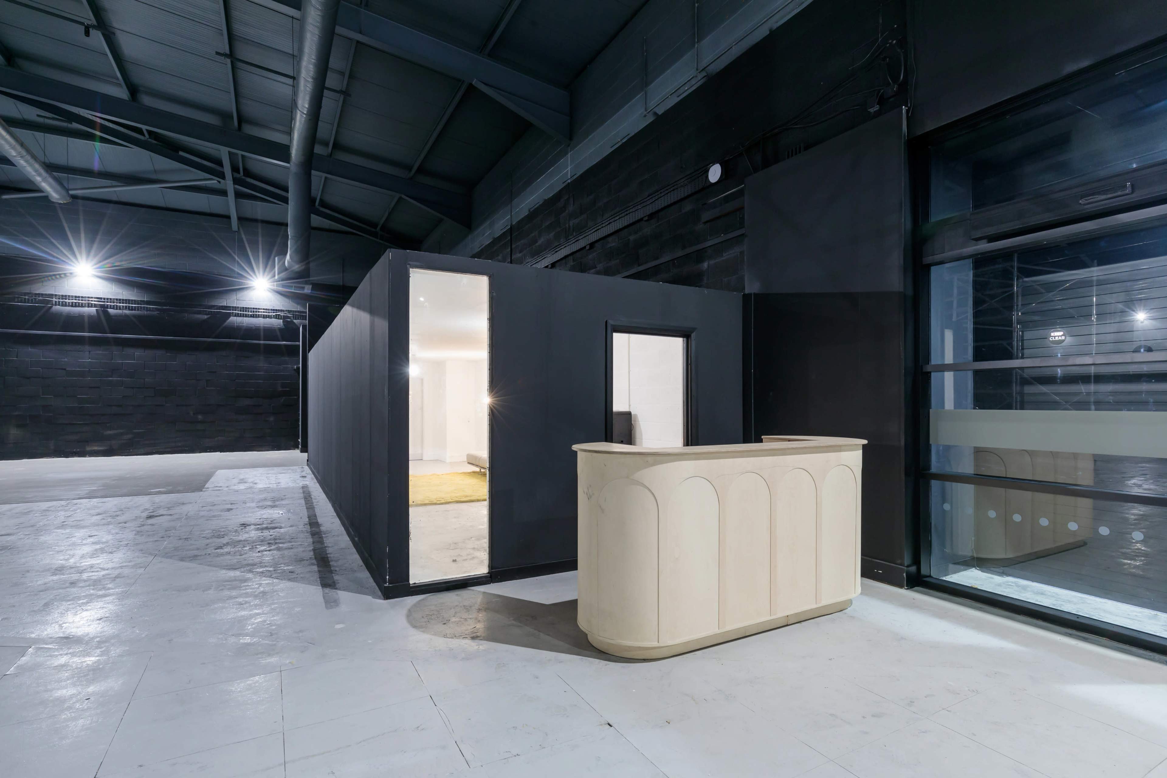 The image shows a modern, minimalist reception area with a light-colored counter and a large, open space featuring a dark wall and industrial ceiling.