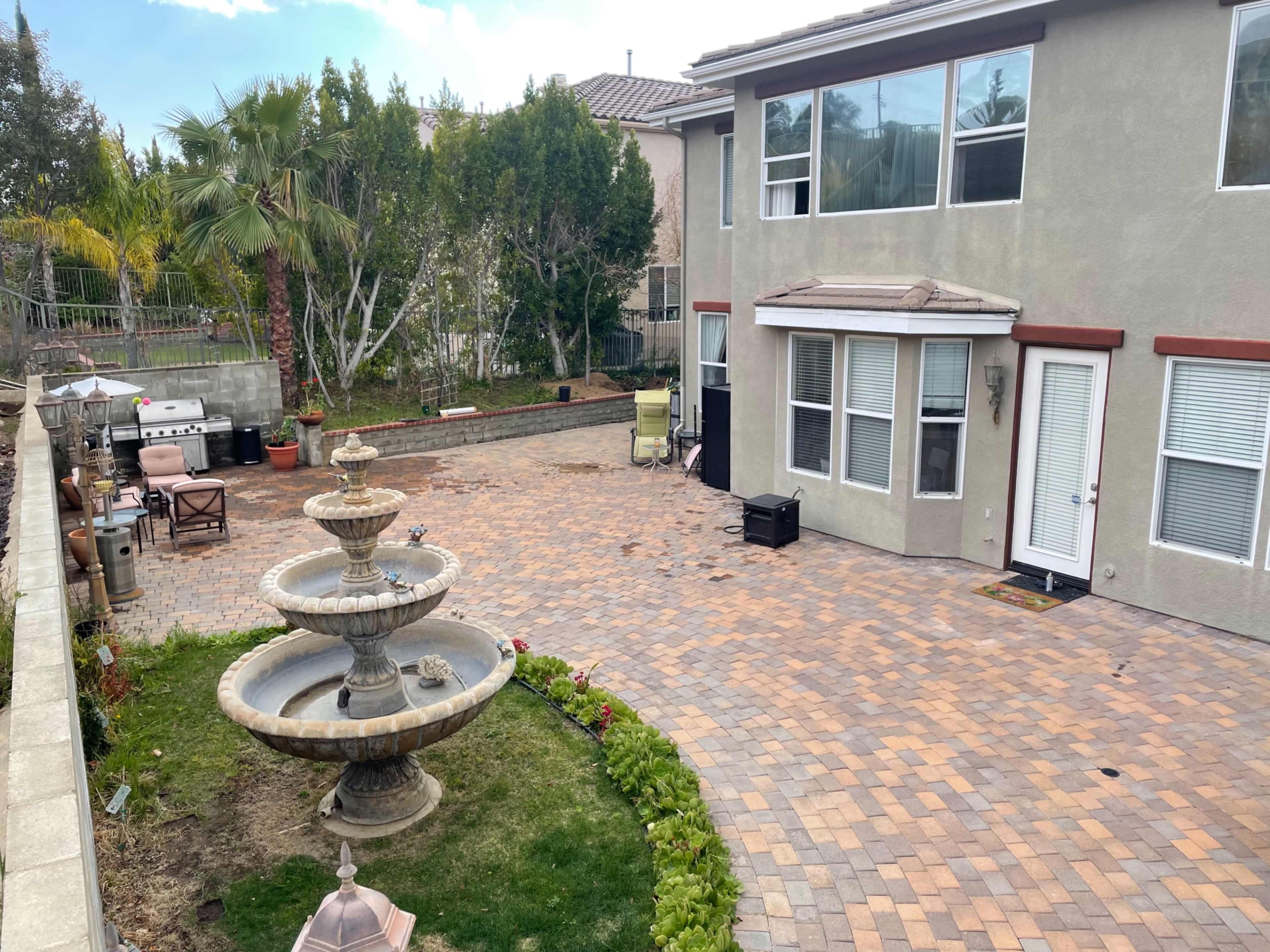 A spacious backyard features a stone patio, a tiered fountain, and well-maintained landscaping with palm trees and bushes.