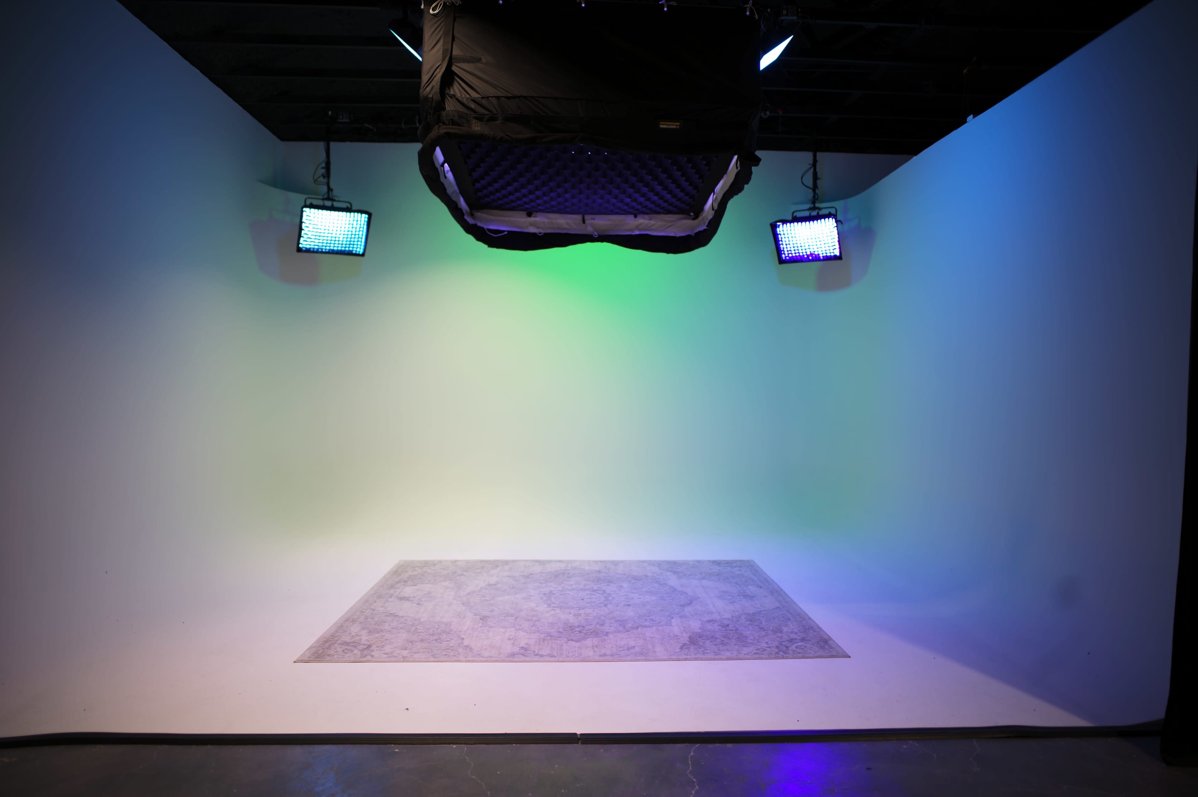 The image shows an empty studio space with a light-colored backdrop, a patterned rug on the floor, and two colorful lights mounted on the walls.