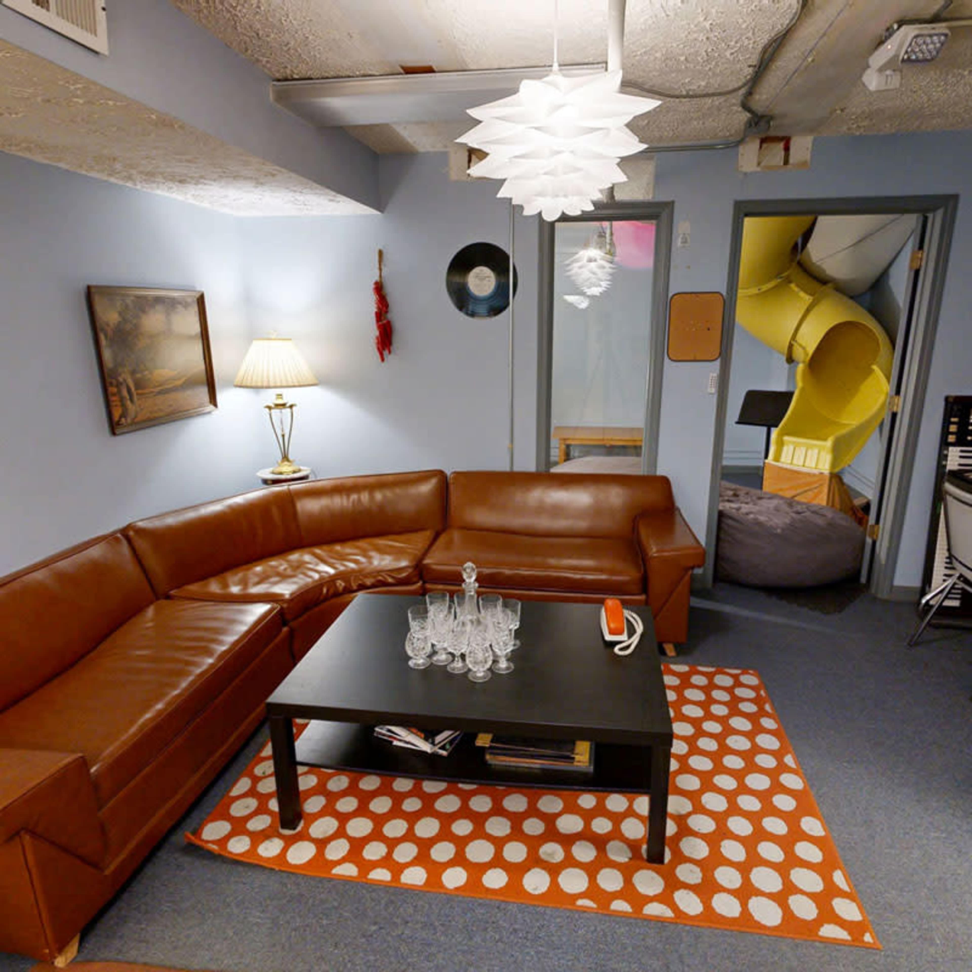 The scene features a cozy basement lounge with a brown sectional sofa, a low black coffee table, a lamp, and a vibrant orange-and-white patterned rug, alongside a bright yellow slide leading to another area.