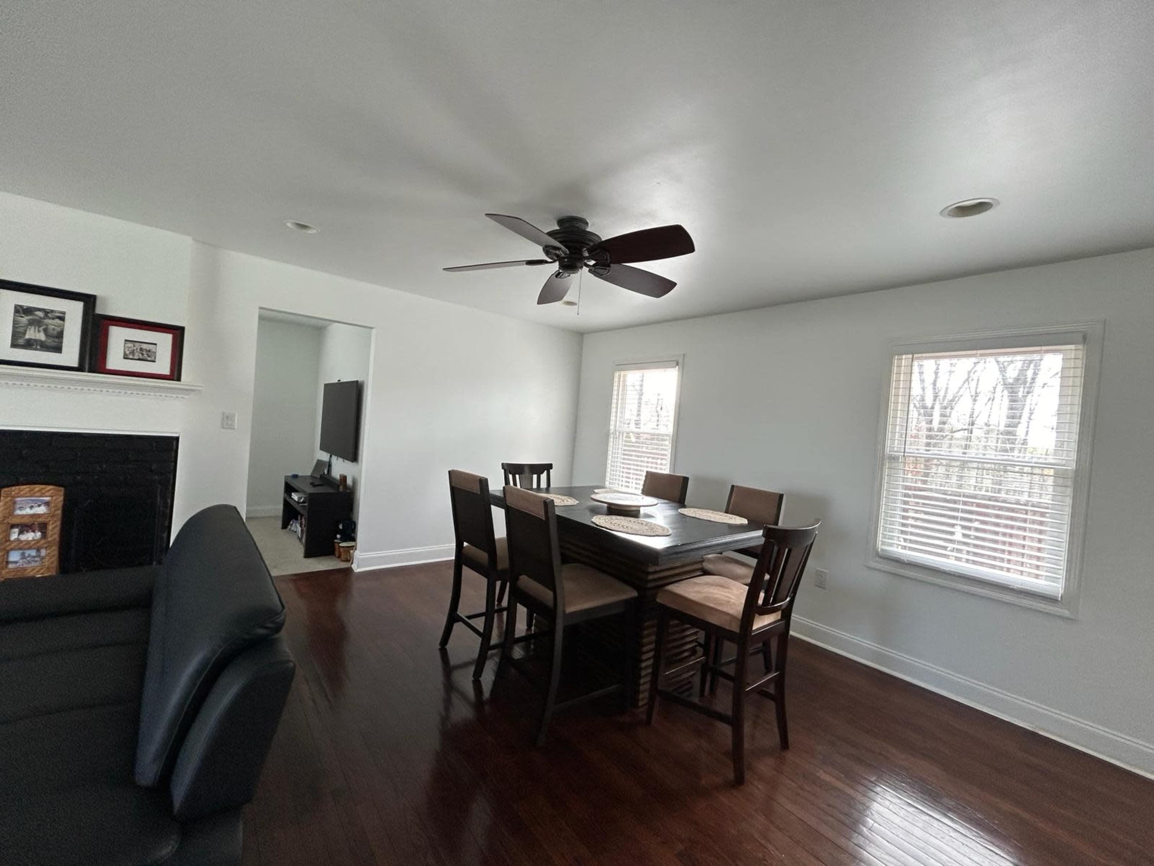 The image shows a dining area with a large table surrounded by eight chairs, a ceiling fan, and two windows allowing natural light to enter.
