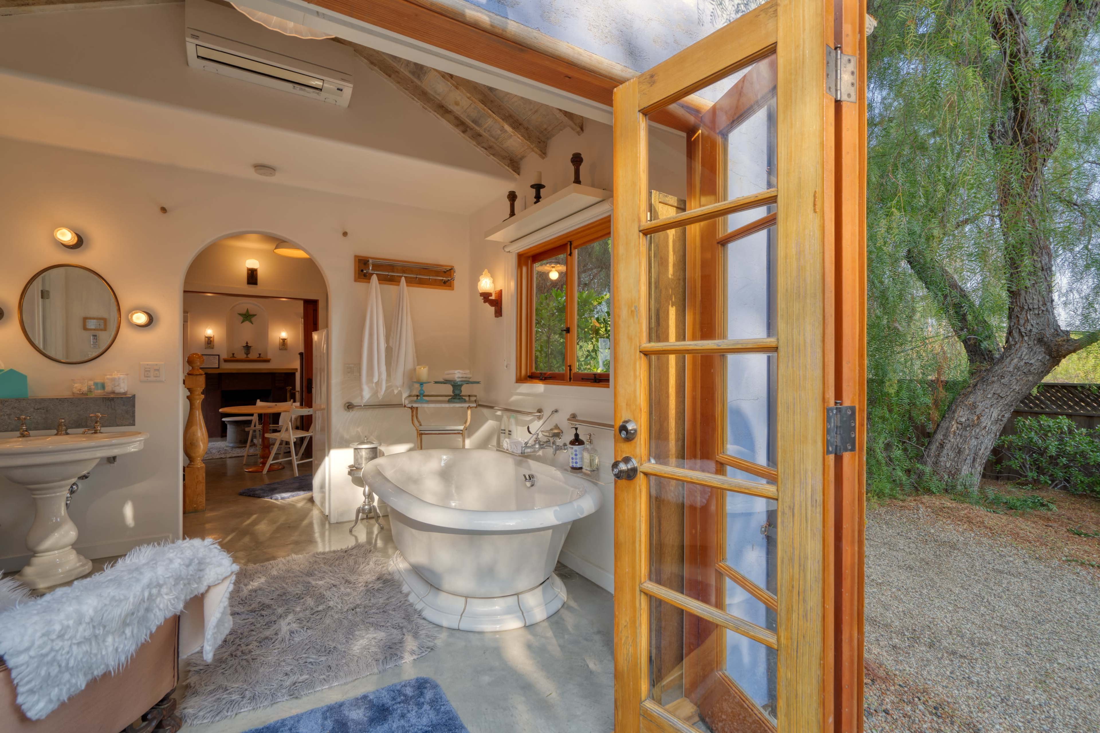 A bright bathroom featuring a freestanding bathtub, wooden door opening to an outdoor area, and a vanity with mirrors and decorative elements.