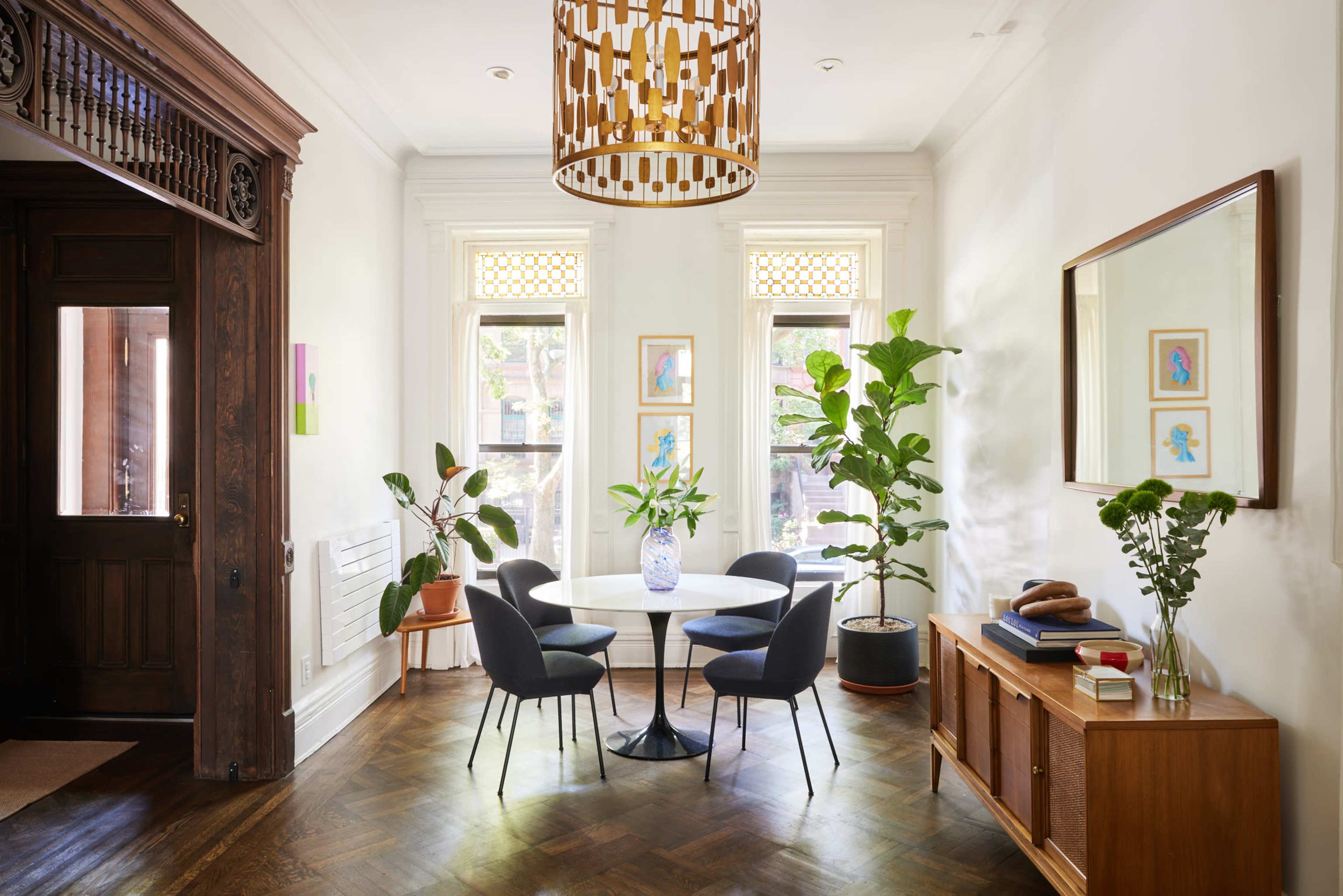 A dining area features a round table surrounded by black chairs, with a large window allowing natural light and decorative plants enhancing the space.