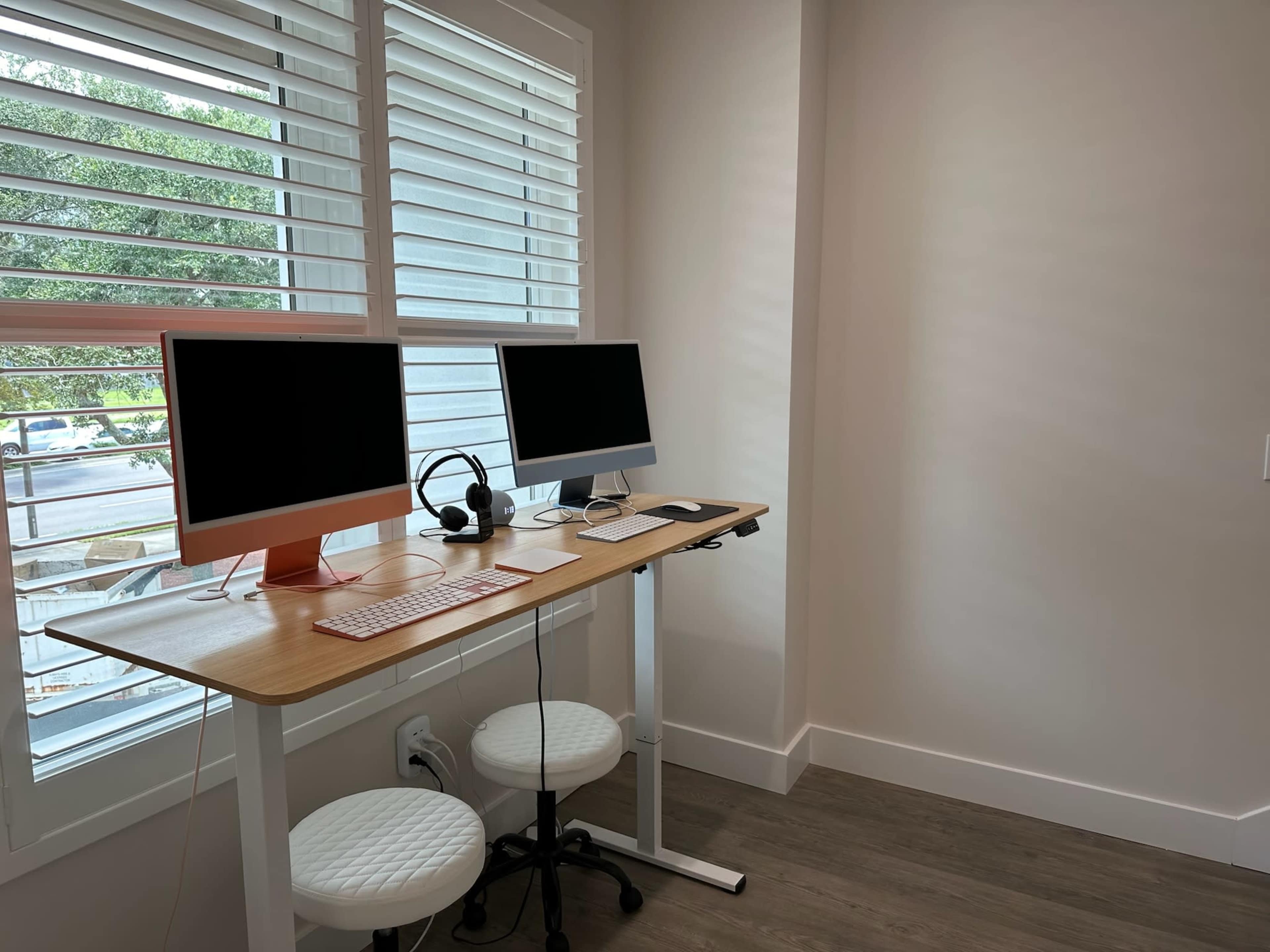 A minimalist workspace with two desktop monitors, a headset, a keyboard, and a mouse on a wooden standing desk next to a window with shutters.