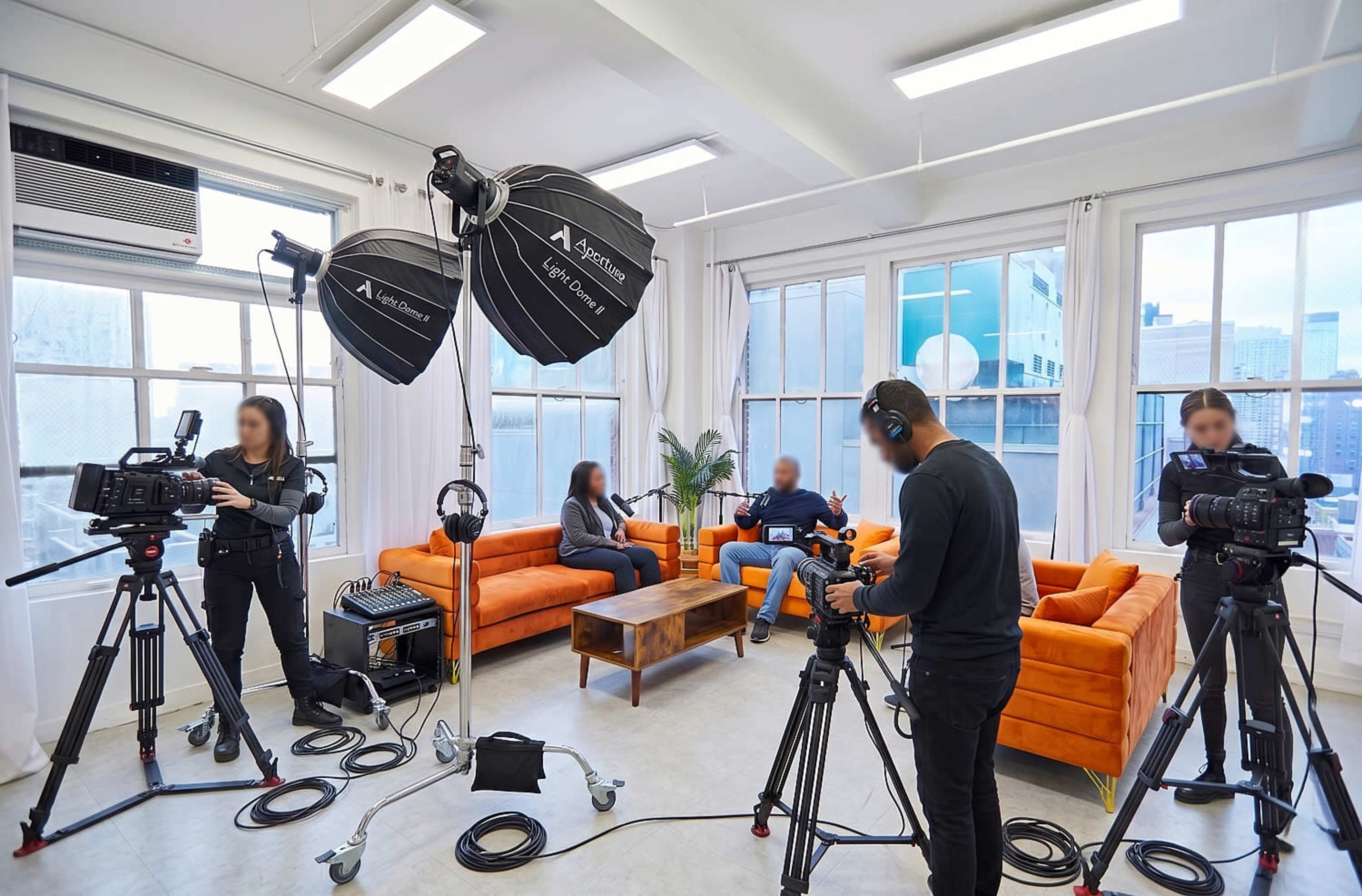 A video production setup features two people seated on orange couches with cameras and lighting equipment arranged around them in a well-lit studio.