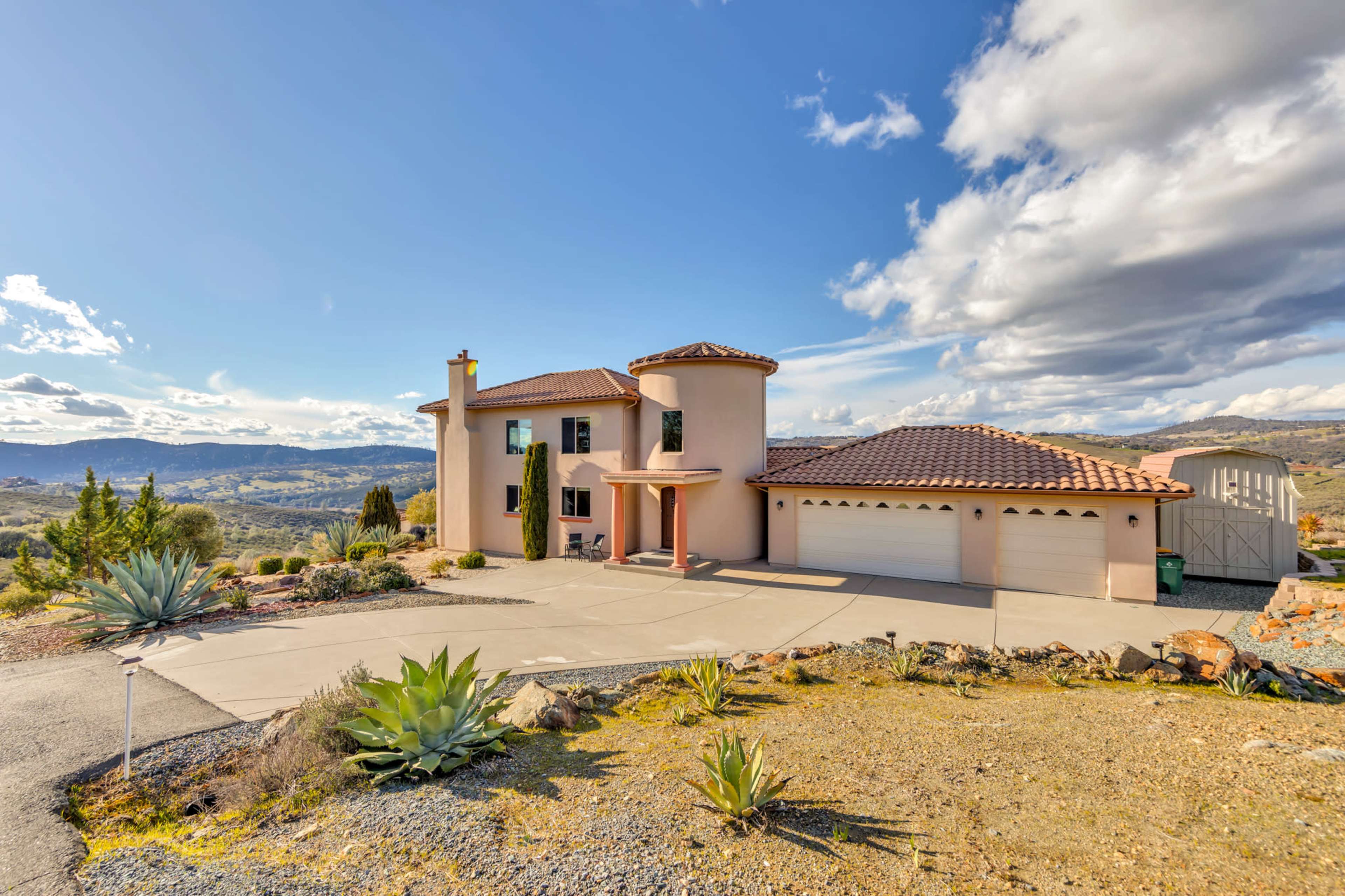 A two-story house with a tiled roof and a three-car garage is situated on a hillside with desert landscaping and distant mountainous views.