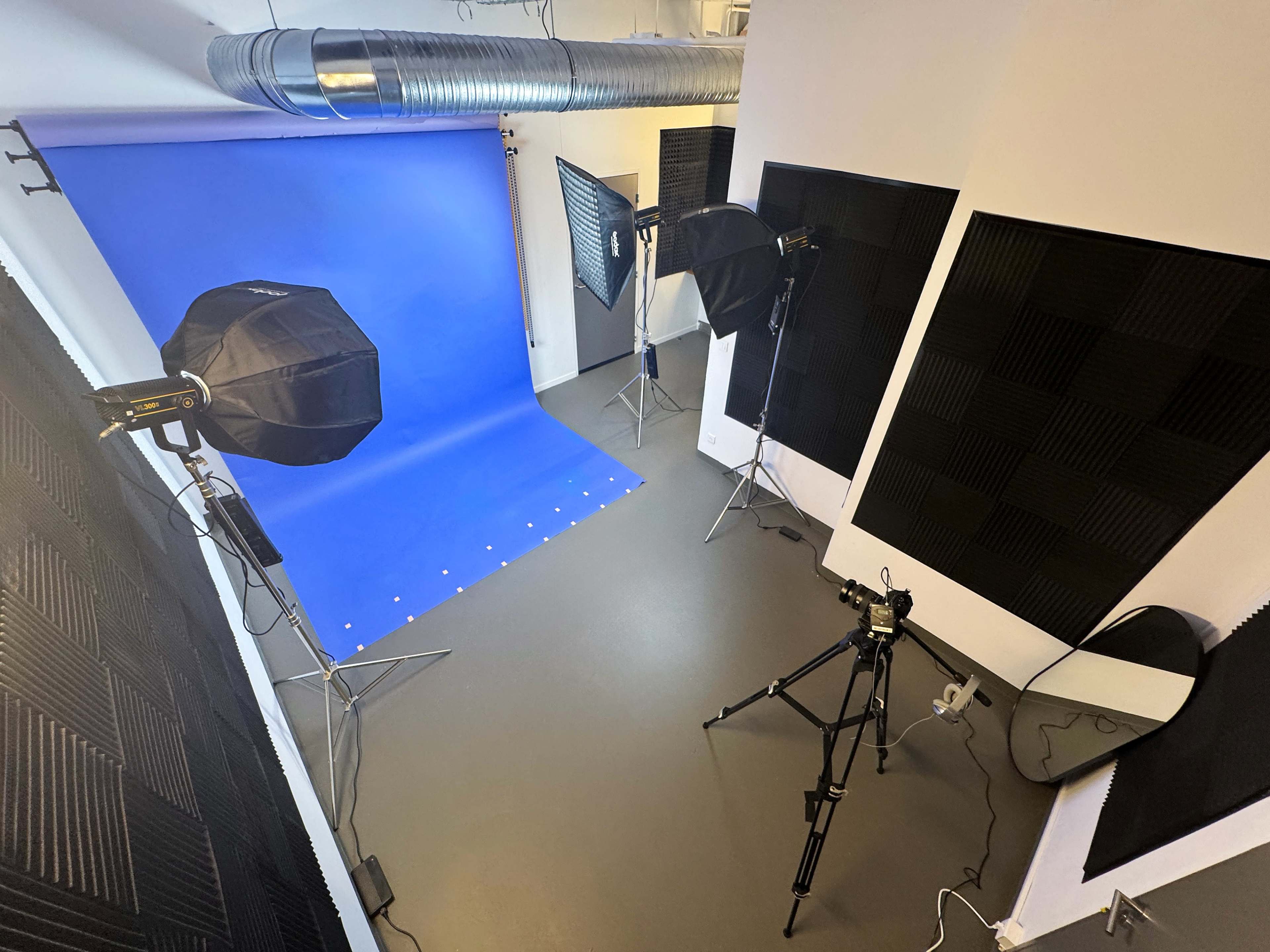 The image shows a photography studio with a blue backdrop, two studio lights, a camera on a tripod, and soundproofing panels on the walls.