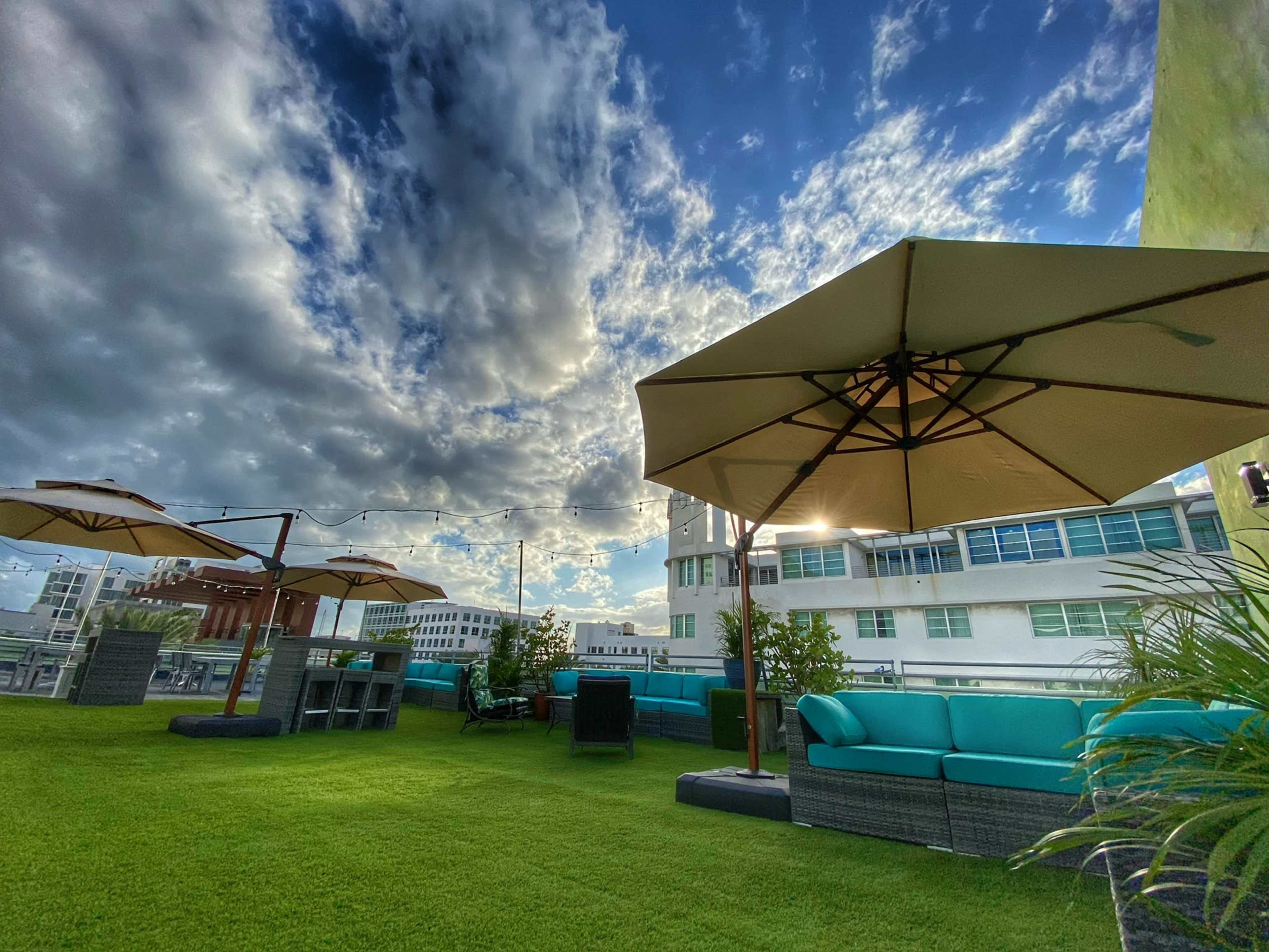 The image shows a rooftop terrace featuring green grass, lounge seating, and umbrellas under a partly cloudy sky.