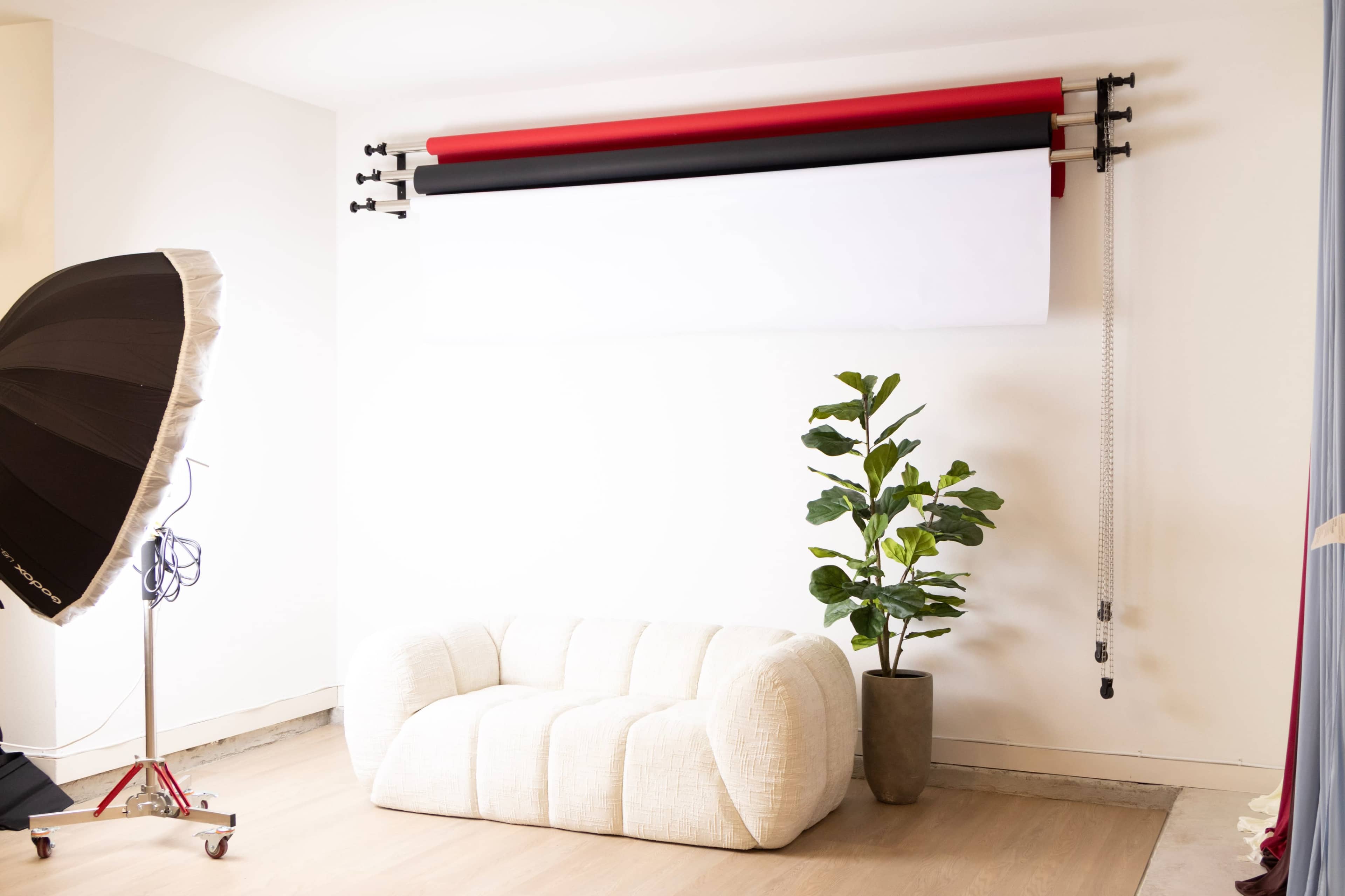 The image shows a minimalist studio setup with a white couch, a potted plant, and three rolled backdrops in red, black, and white mounted on a wall.