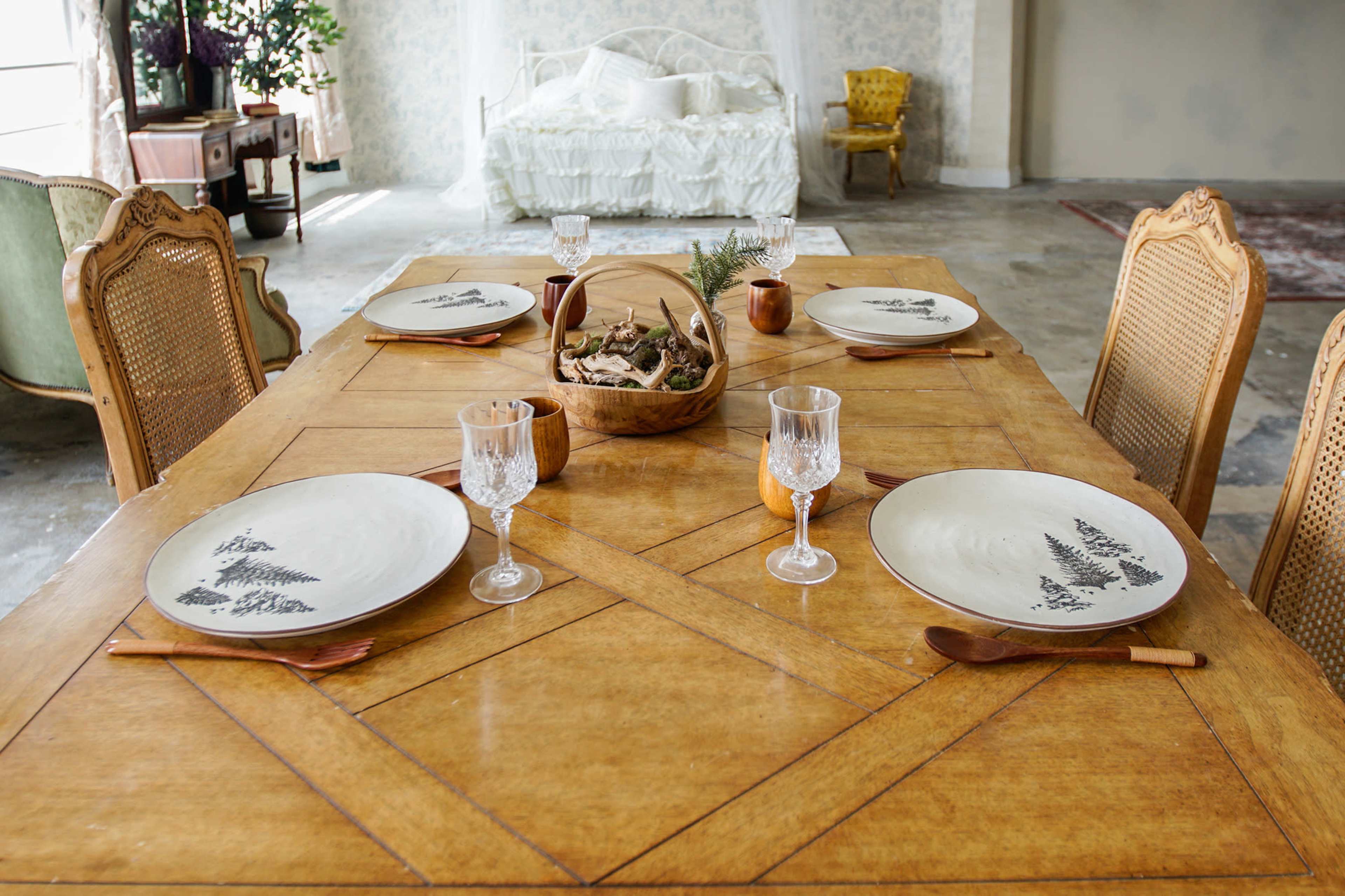A wooden dining table is set with four plates, crystal glasses, and a decorative bowl filled with natural materials, surrounded by a light-filled room with vintage furniture.