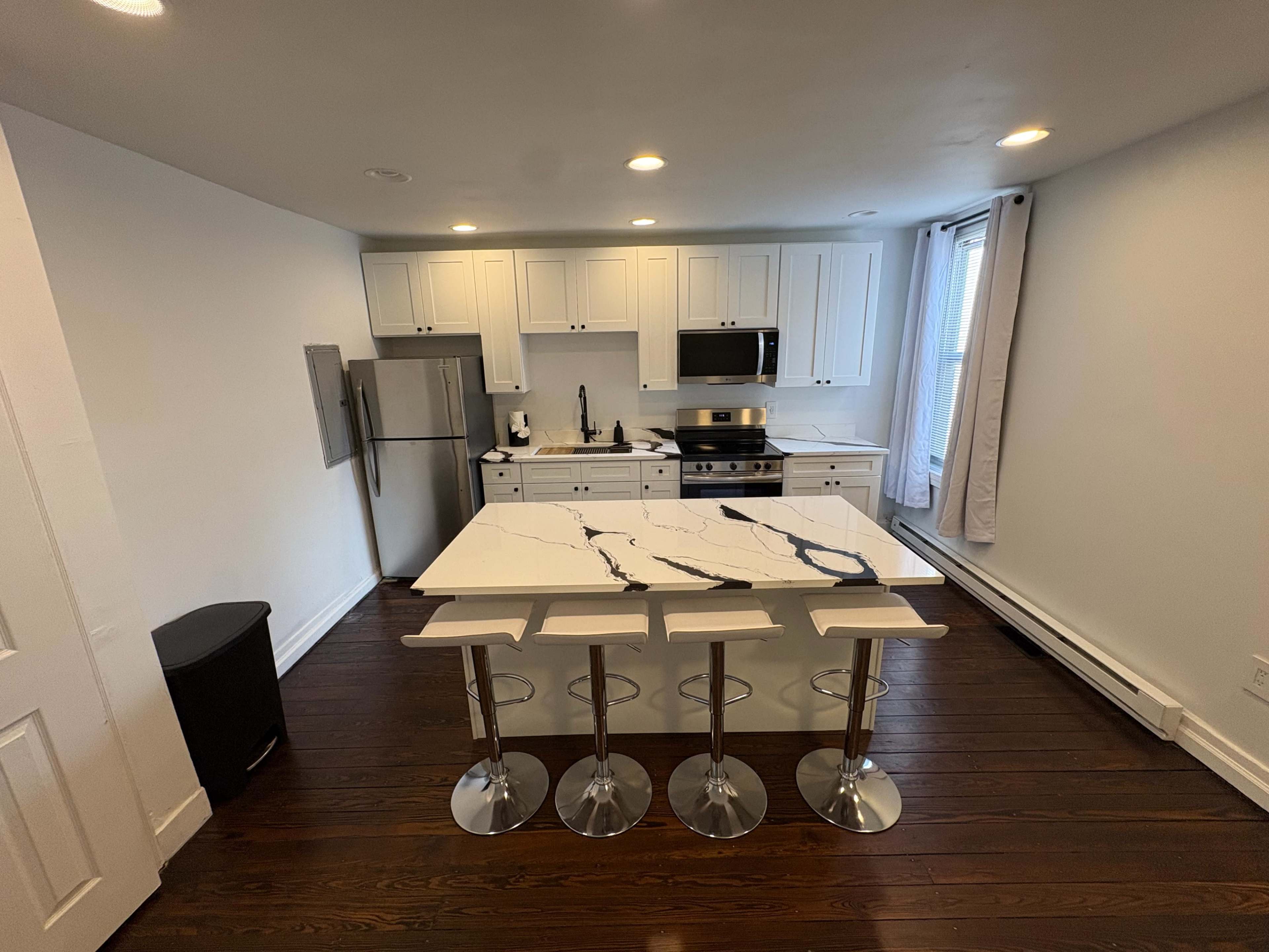The image shows a modern kitchen with white cabinetry, stainless steel appliances, and a large island featuring a marble-patterned countertop and three metal stools.
